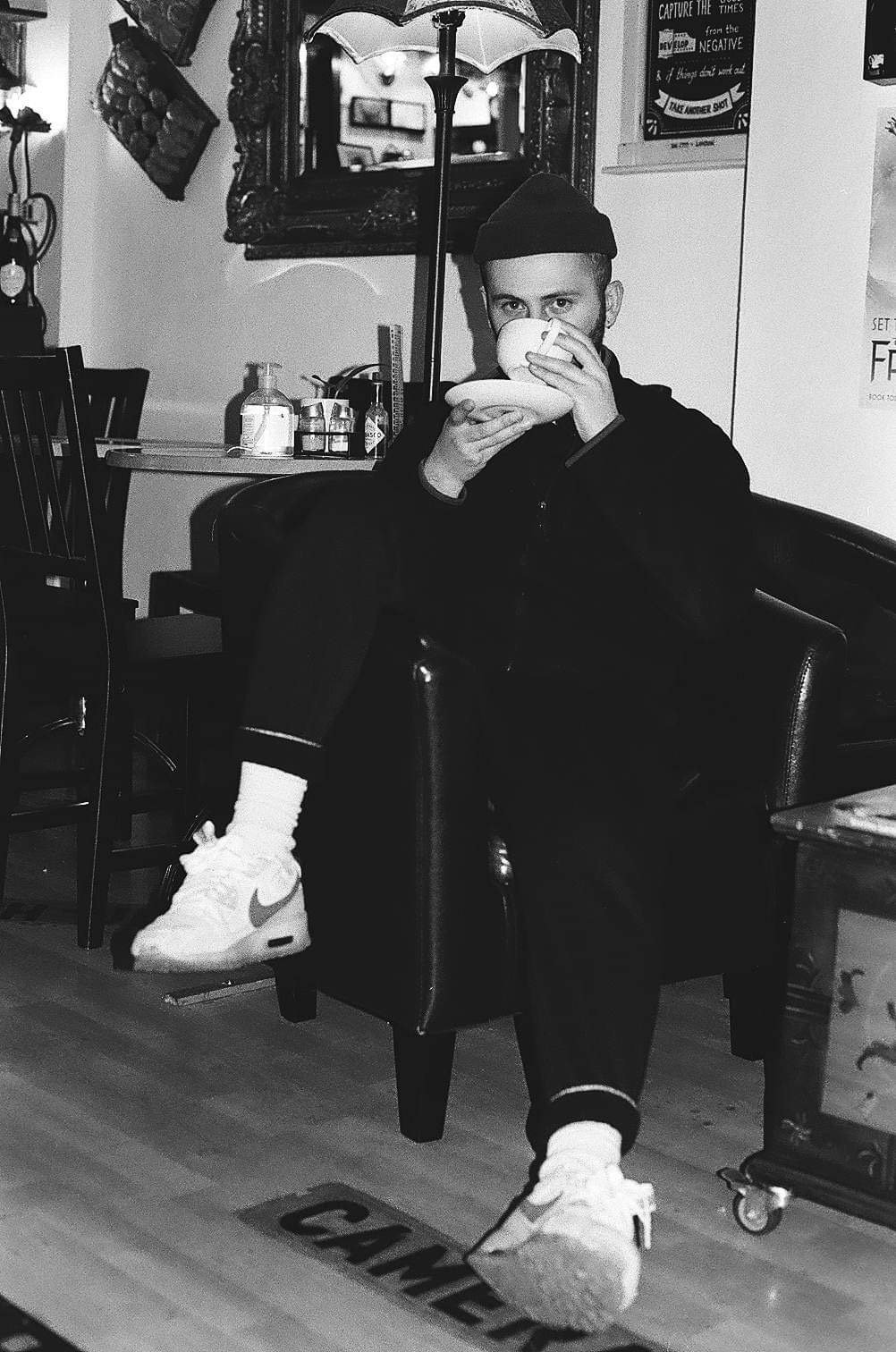 A man sitting on a leather armchair inside a café, drinking from a teacup and saucer. He is wearing a black hoodie, white sneakers, and a beanie. His right foot rests on a sign on the floor that reads 'CANCELED'. The background includes a mirror, a small table with a hand sanitizer, a remote control, and some bottles, with wall decorations and a bulletin board.