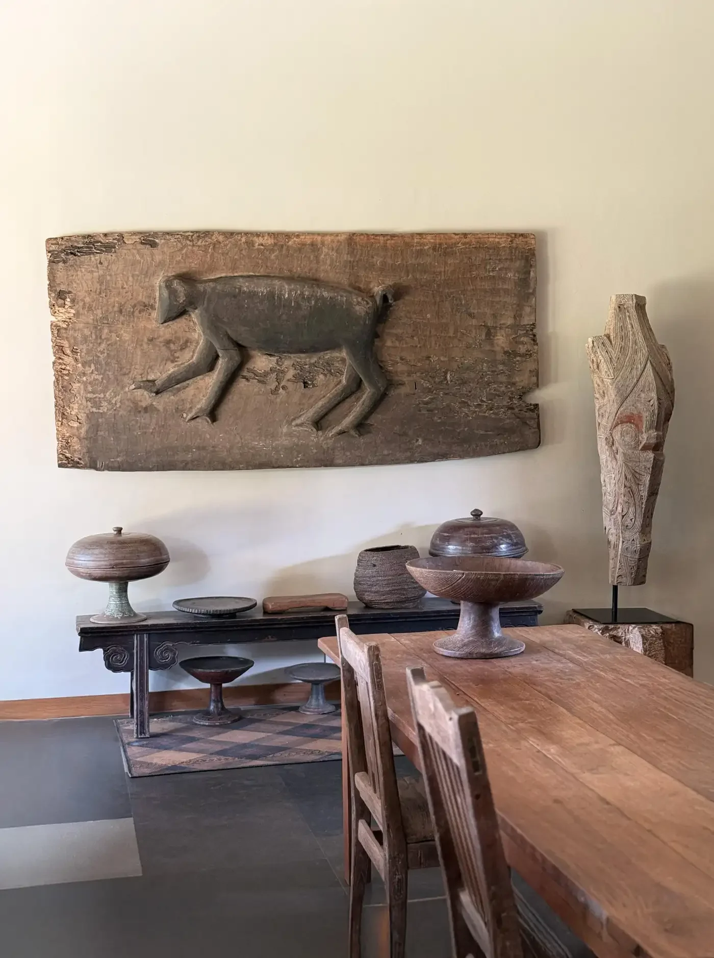 Room with wooden furniture and wall art, featuring a carved pig on a large wooden panel, a wooden table, chairs, and various wooden decorative objects.