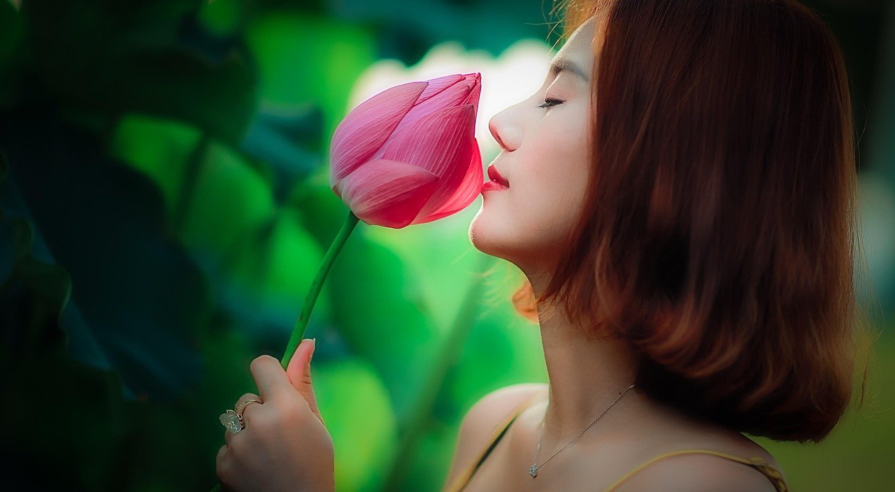 A woman with shoulder-length brown hair and fair skin smelling a pink flower outdoors, with a blurred green background.