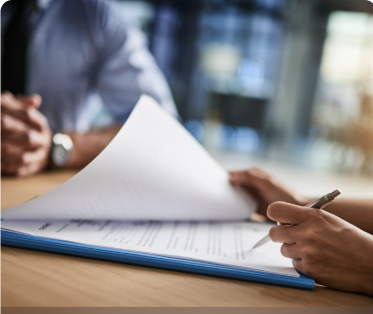 Close-up of two people reviewing a document at a table, one person holding a pen and the other flipping through papers, with a blurred office background.