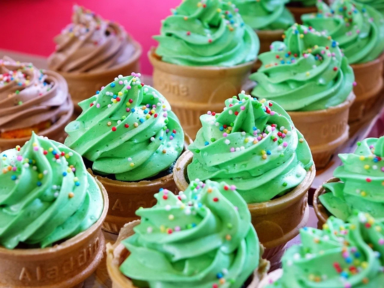 Cup cakes with bright green frosting and colorful sprinkles against a pink background.