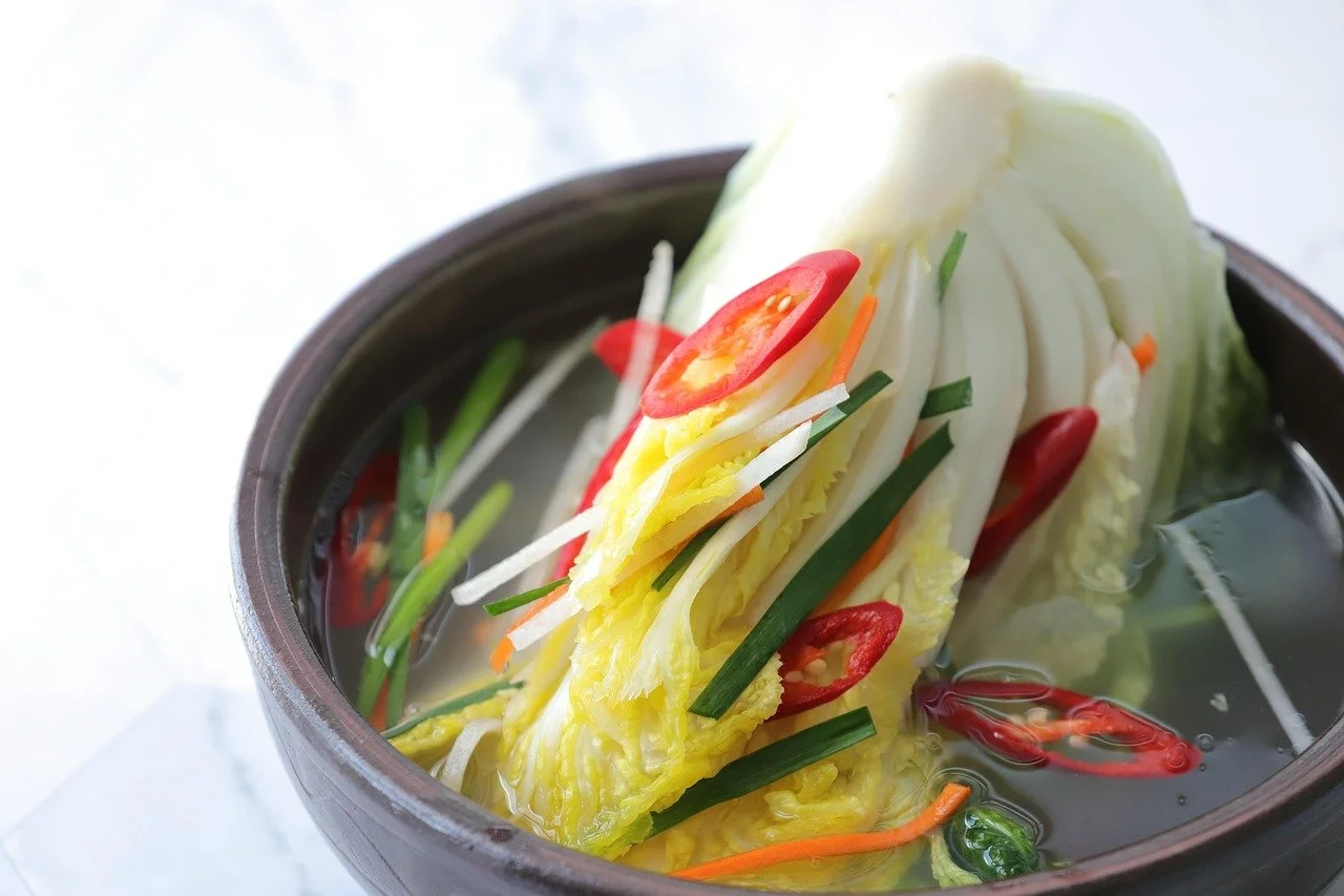 A bowl of hot Asian-style soup with kimchi made from  leafy napa cabbage, sliced red chili peppers, shredded carrots, and chopped green onions, floating in a clear broth.