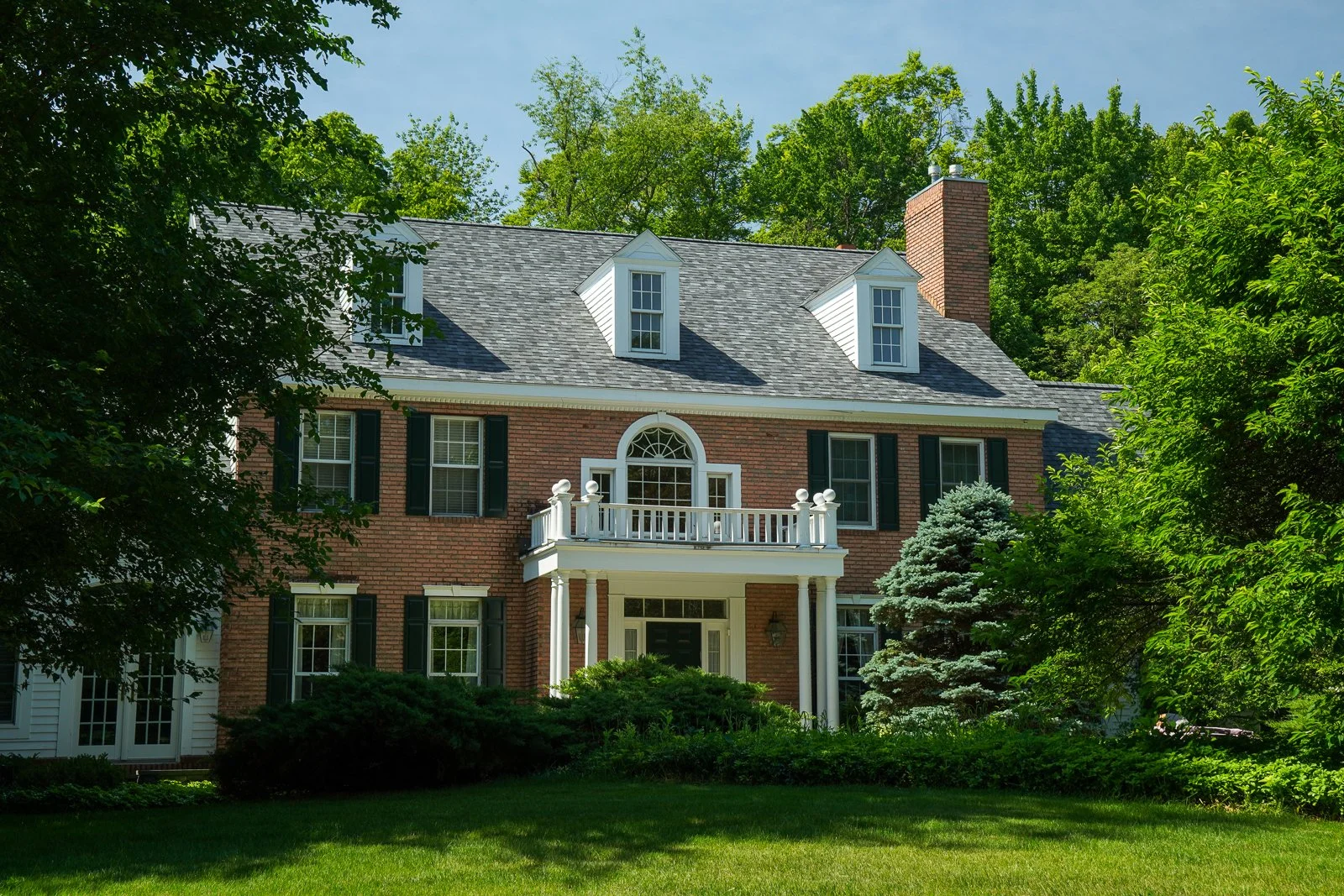A large brick house with a gray shingled roof, dormer windows, and a balcony above the front porch, surrounded by green trees and bushes under a partly cloudy sky.