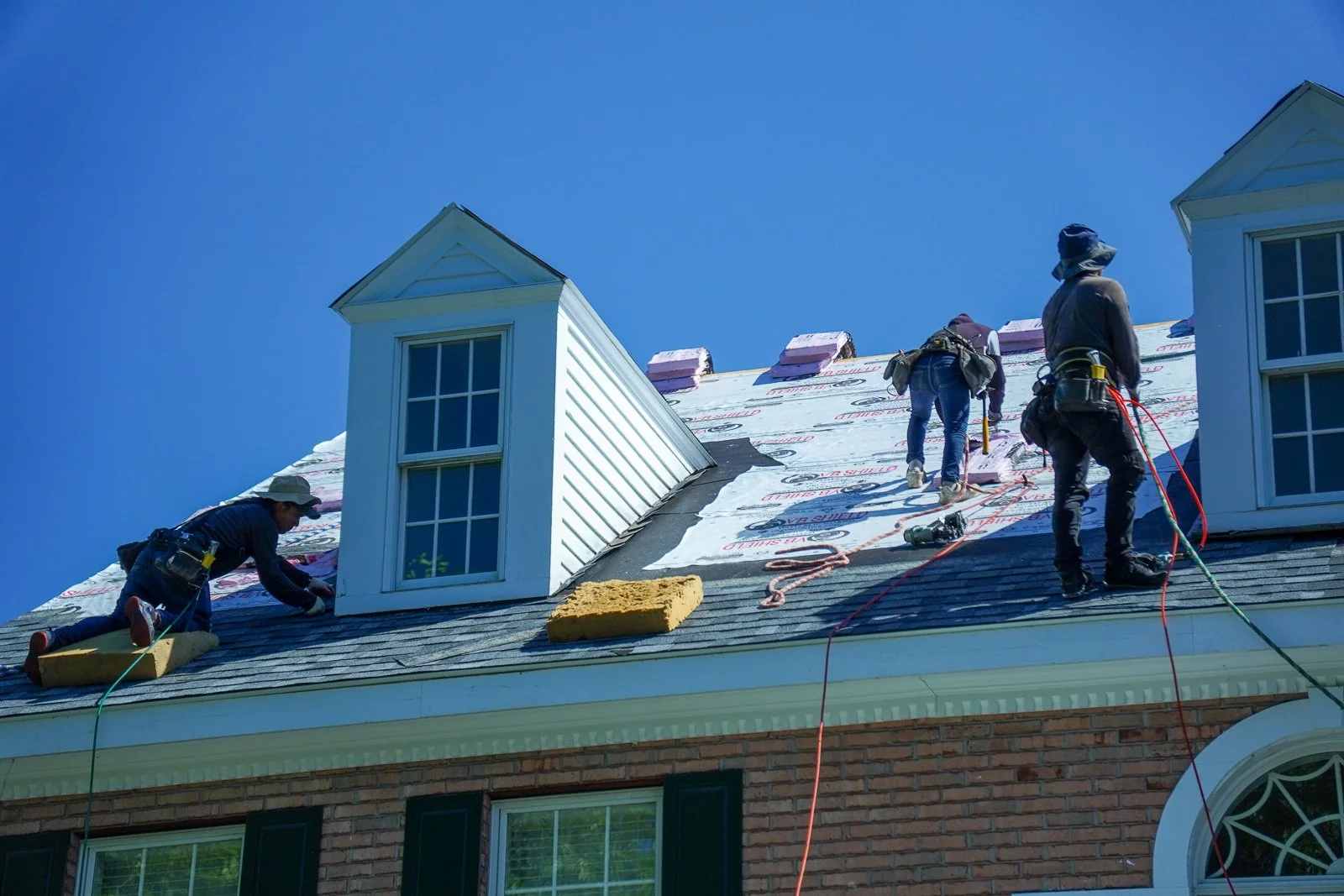 Workers installing or repairing roofing on a house, some on the roof and others preparing tools and materials.