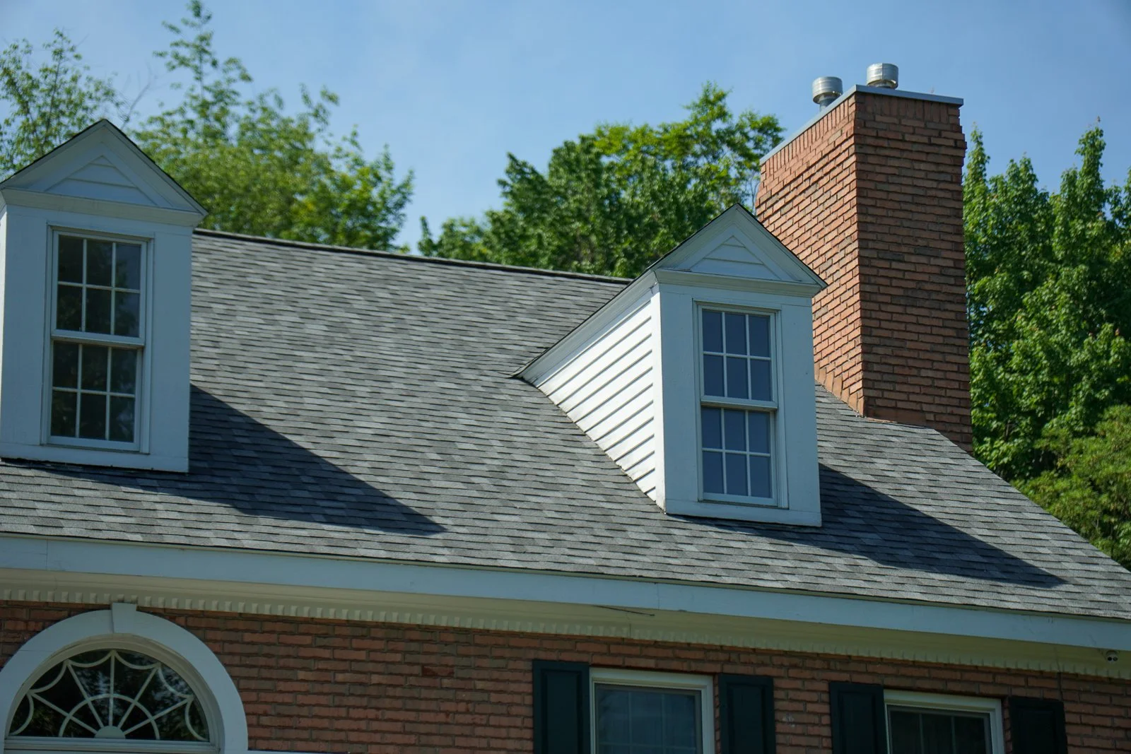 Close-up of the roof of a house with two dormer windows, a brick chimney with vents, and green trees in the background under a blue sky.