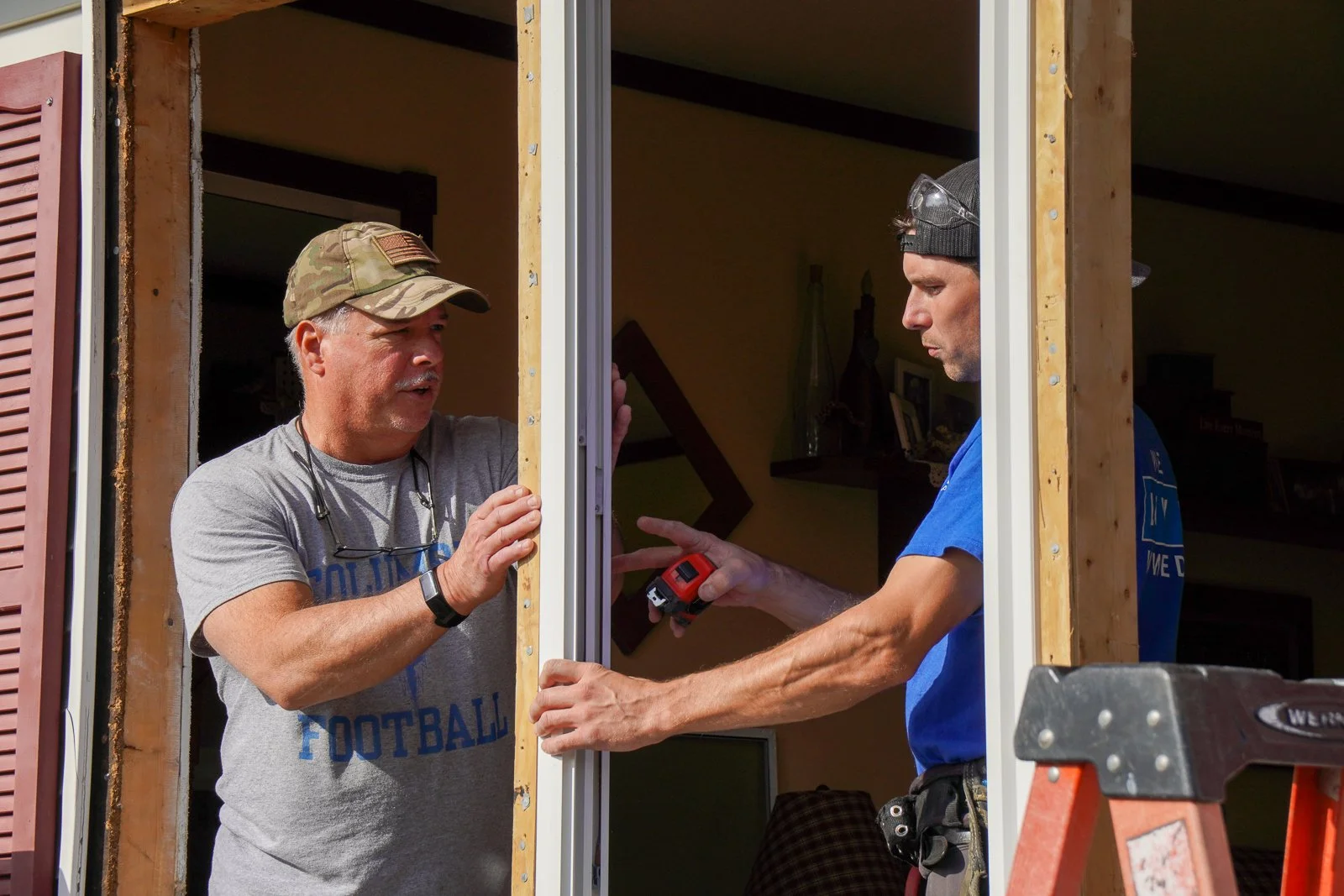 Two men working on a home renovation, installing or fixing a window. One man is wearing a camouflage cap and a grey T-shirt, the other is in a blue shirt with safety glasses on his head. They are inside a partially constructed window frame.