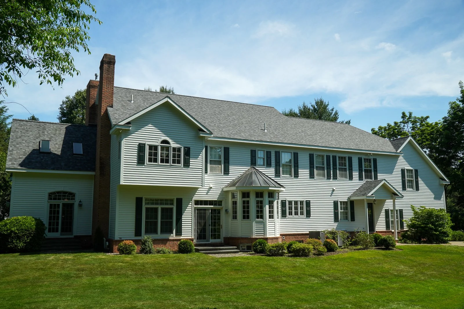 Large white house with black shutters, multiple windows, and a brick chimney, surrounded by a well-kept lawn and trees, under a partly cloudy sky.