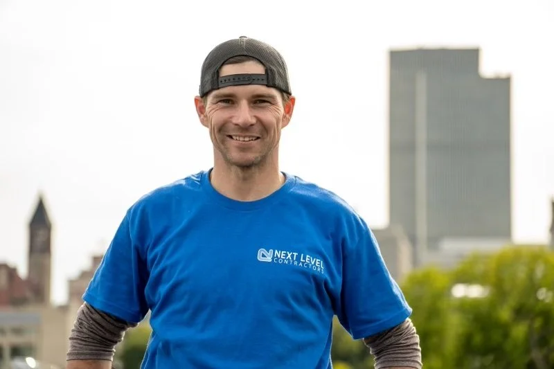 A smiling man wearing a gray baseball cap backwards and a blue T-shirt with 'Next Level Contractors' logo, standing outdoors in front of a cityscape with buildings and greenery.