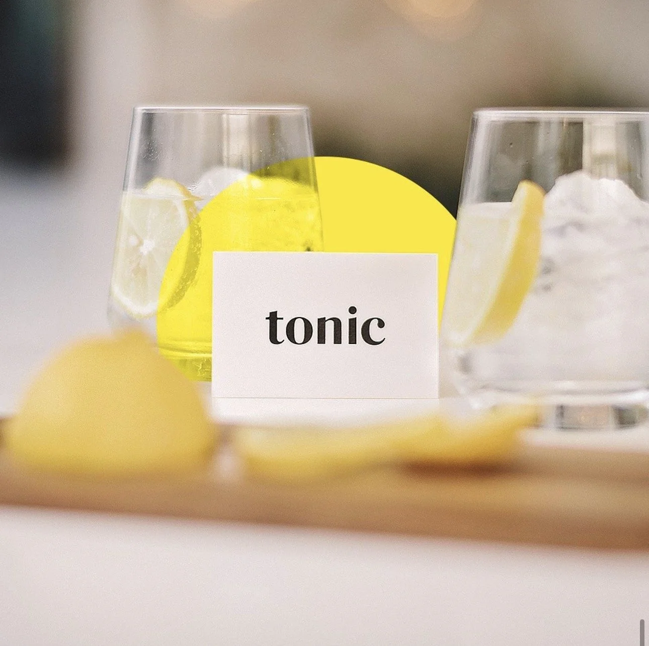 Two glasses with ice, lemon slices, and a lemon wedge on a coaster, labeled 'tonic' on a white card with a yellow background in the center.