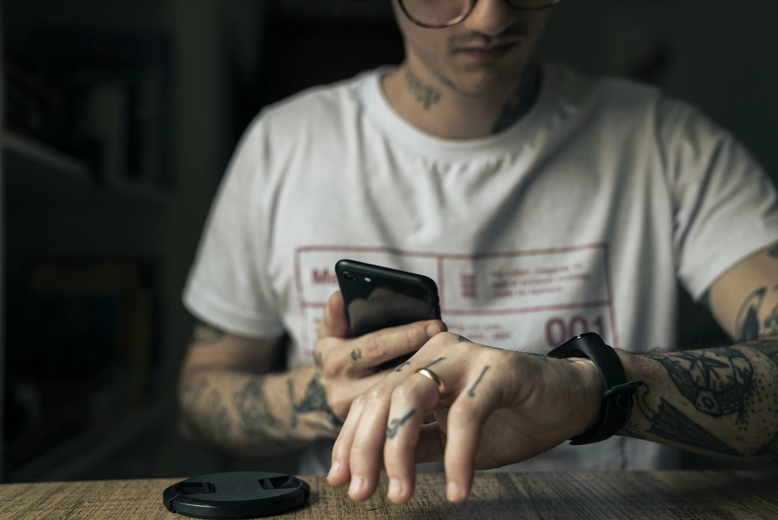 Person with tattoos wearing a white t-shirt, holding a smartphone, and adjusting a smartwatch. A camera lens cap is on the wooden table.