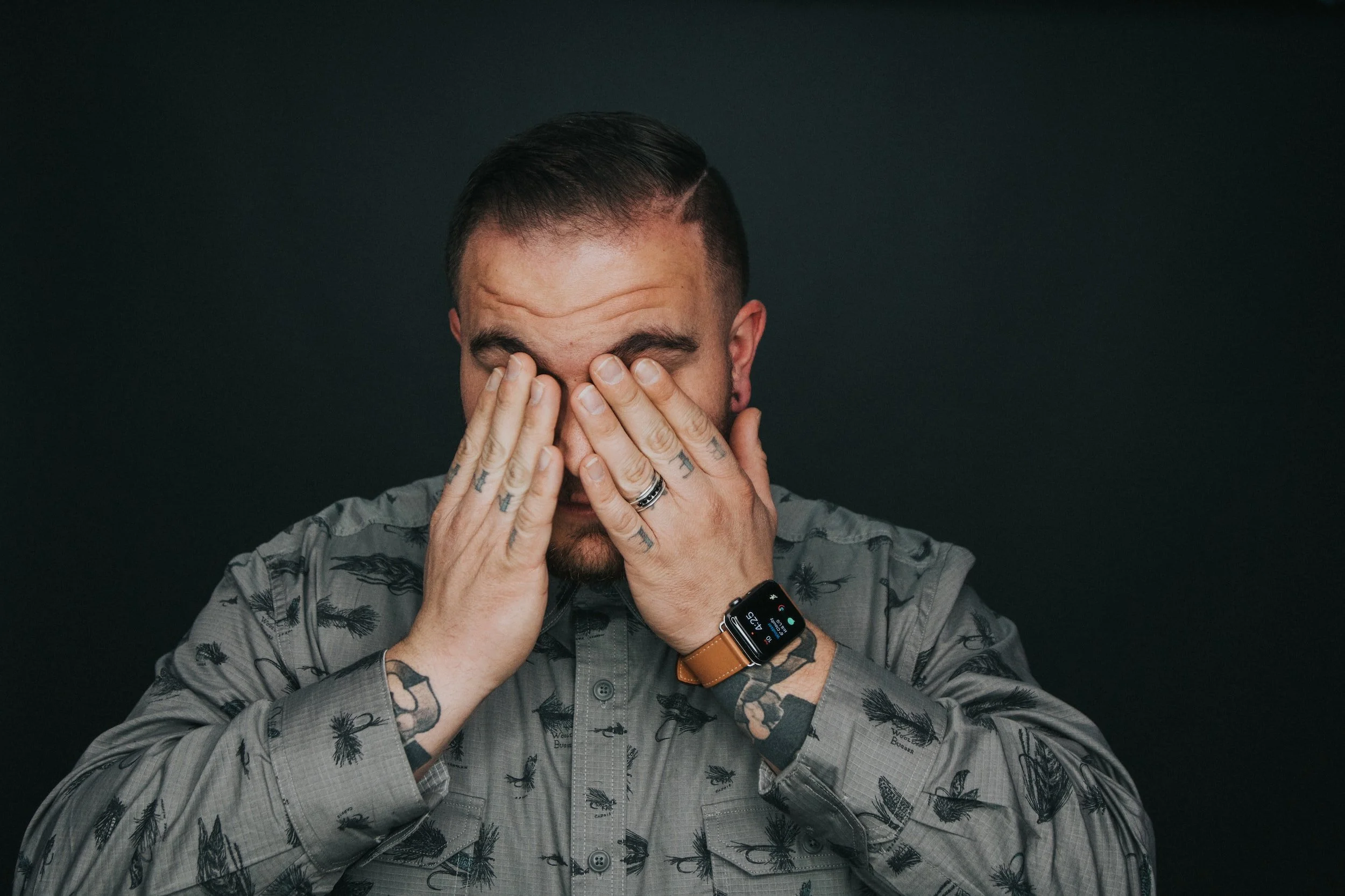 Man in gray patterned shirt covering his eyes with hands, showcasing tattoos on fingers and wearing a smartwatch.