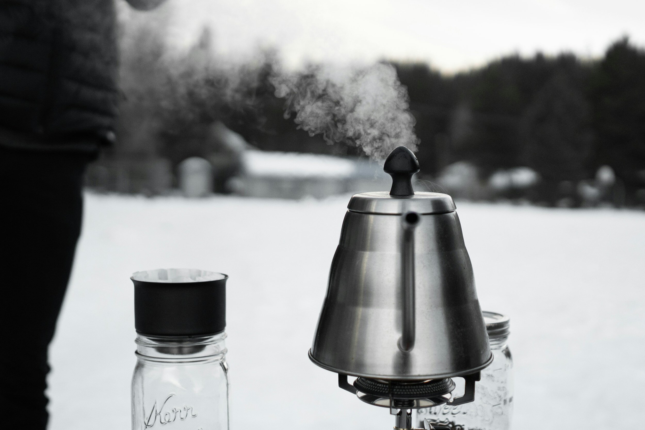 Steam rising from a stainless steel kettle beside glass jars, outdoor setting with snowy background.