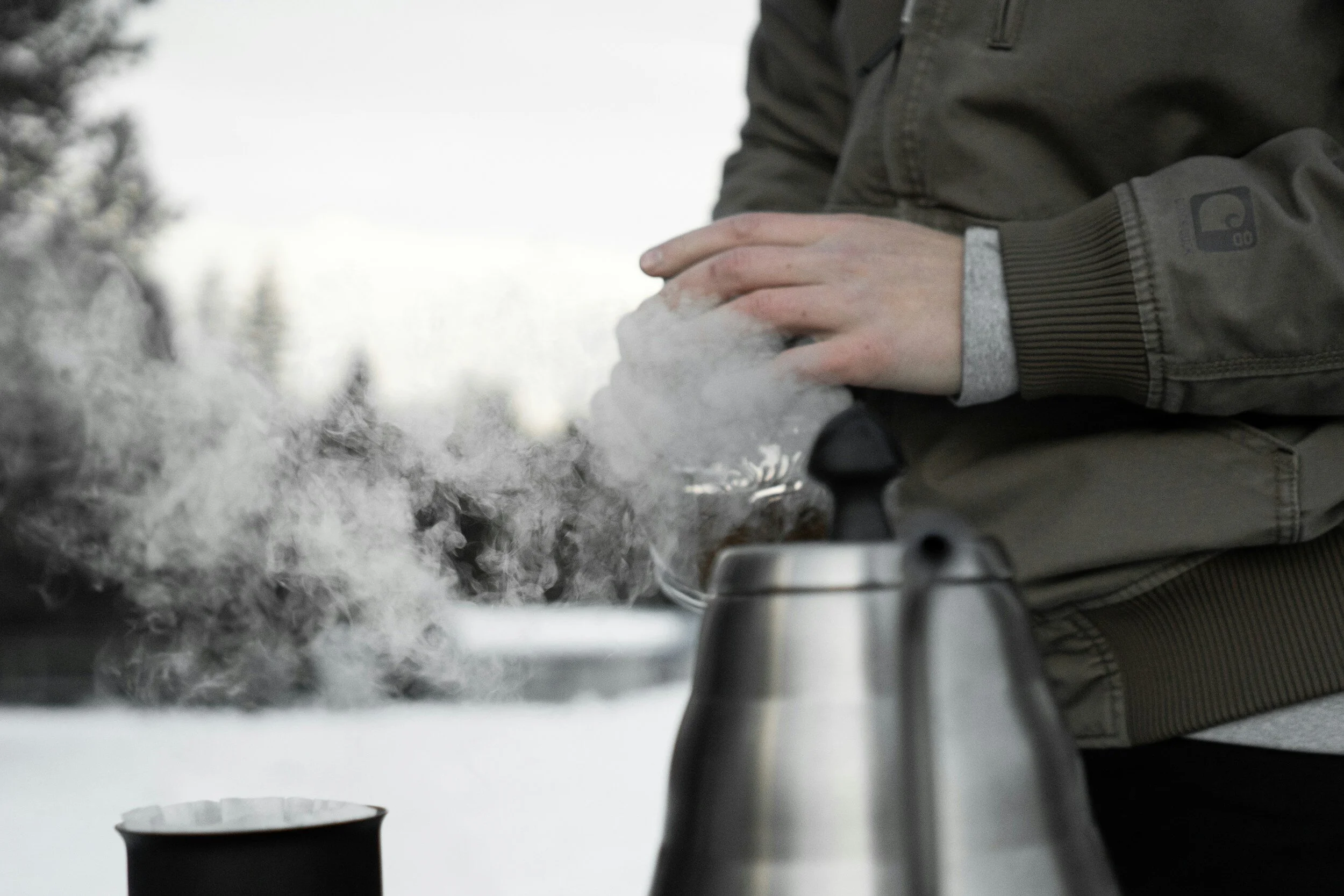 Person pouring hot water from a silver kettle with steam, outdoors.