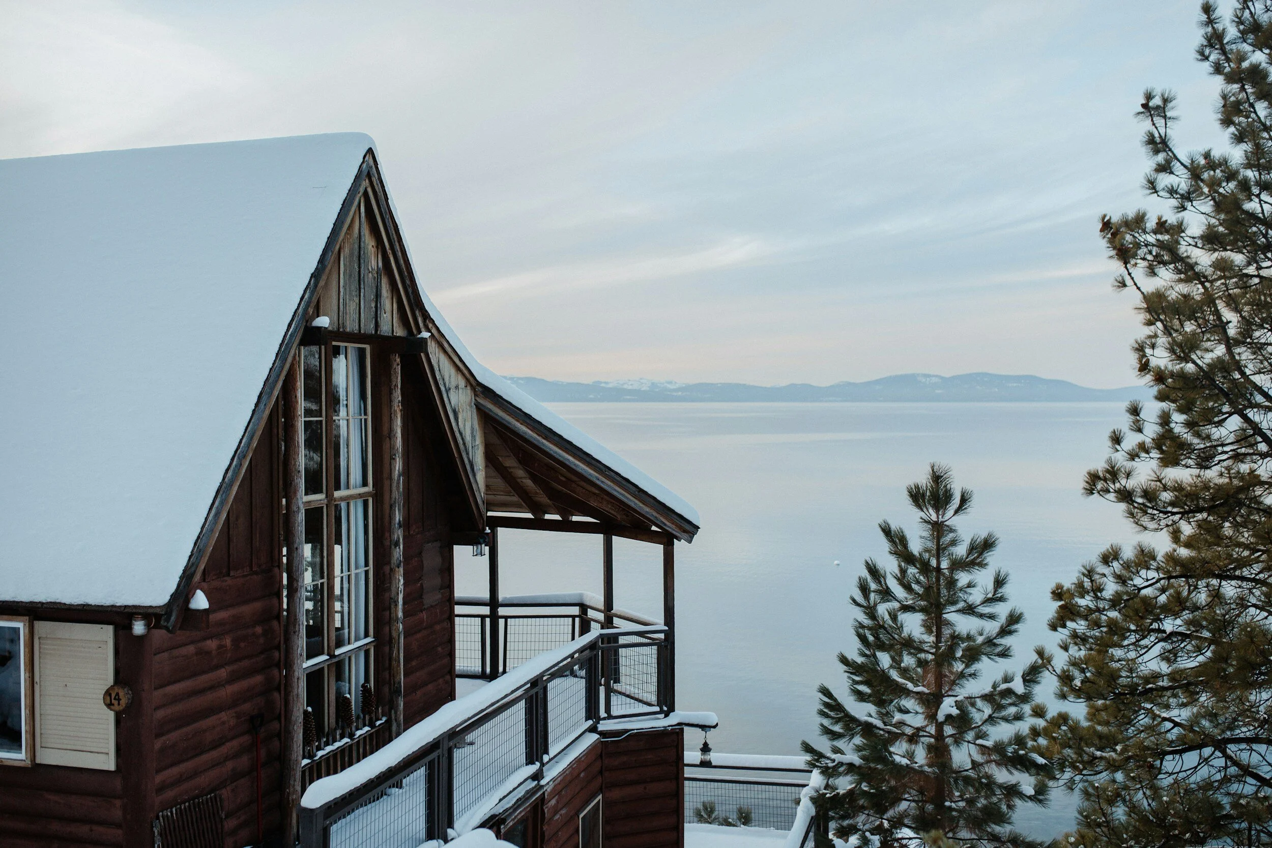 A snow-covered wooden cabin with large windows overlooking a serene lake with distant mountains under a clear sky. Pine trees surround the cabin.