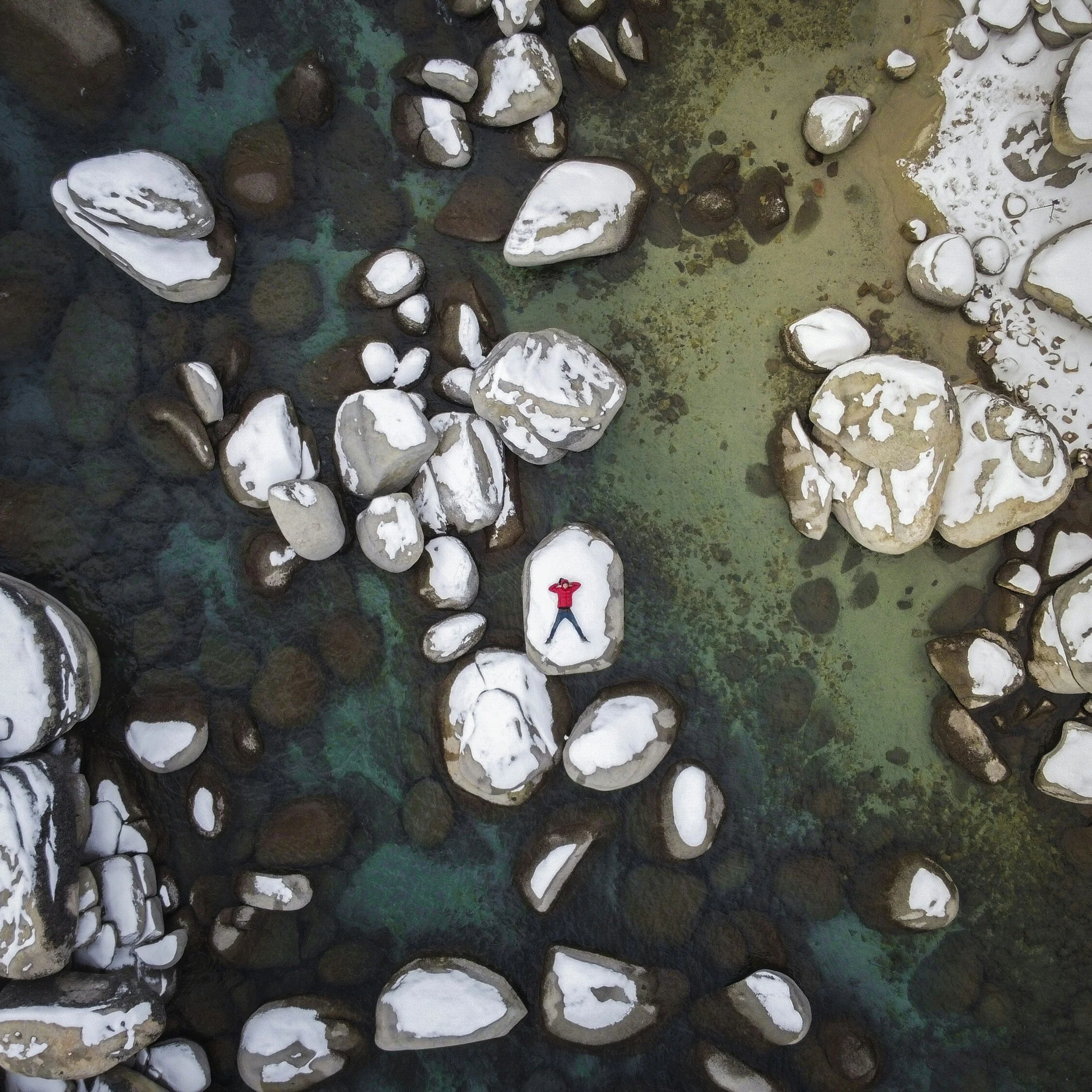 Aerial view of a person in a red jacket lying on a snow-covered rock surrounded by other snow-dusted rocks in shallow, clear water.