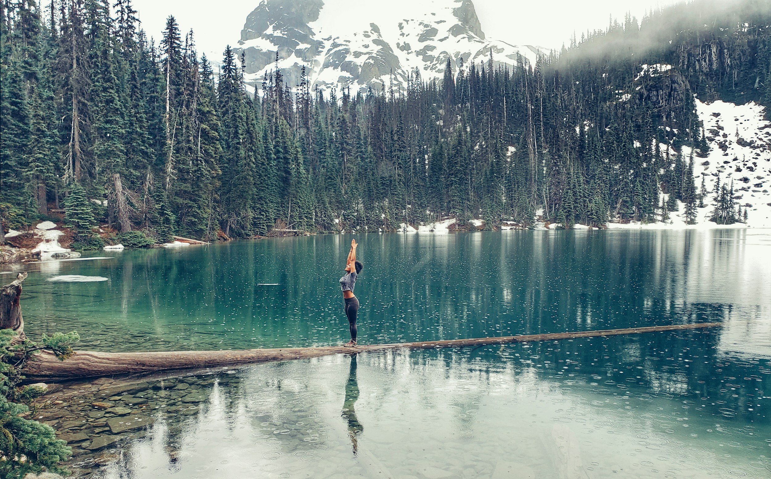 Person doing yoga on a log over a turquoise lake surrounded by pine trees and snowy mountains.