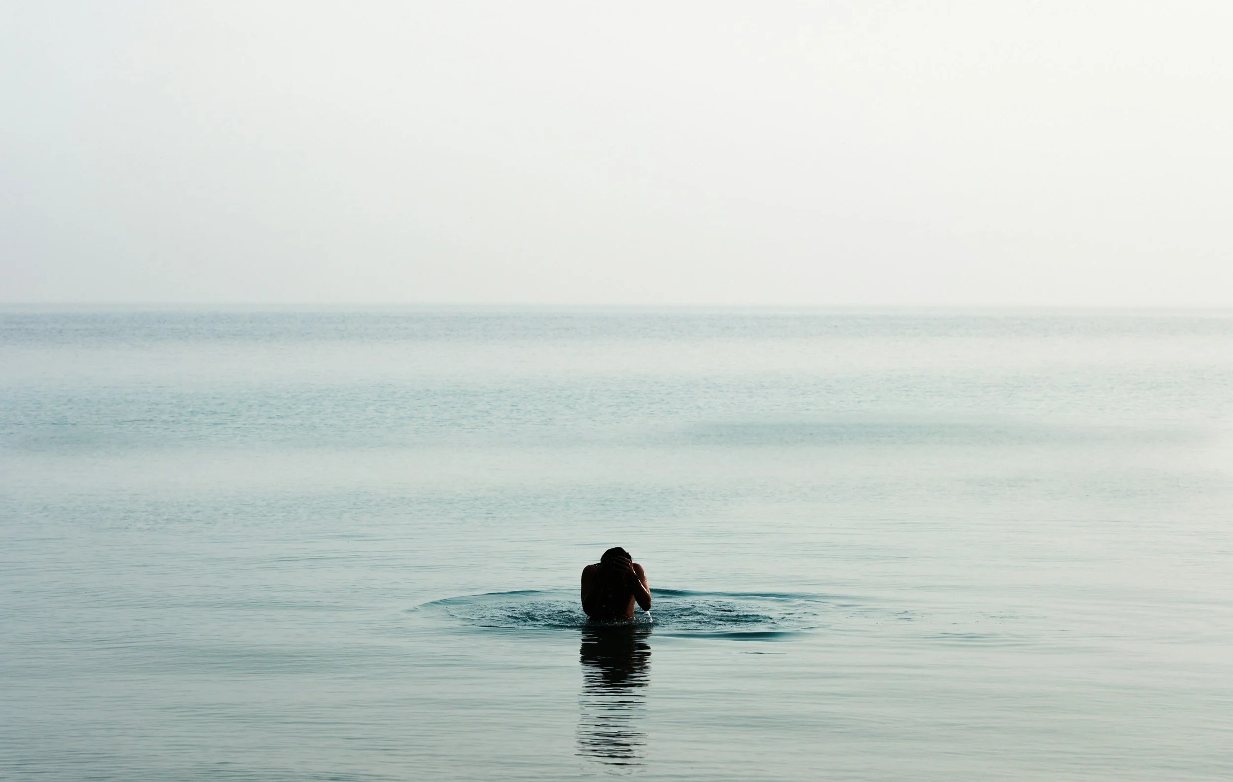 Person standing in calm water at sea, facing away, with a blurred horizon in the background.