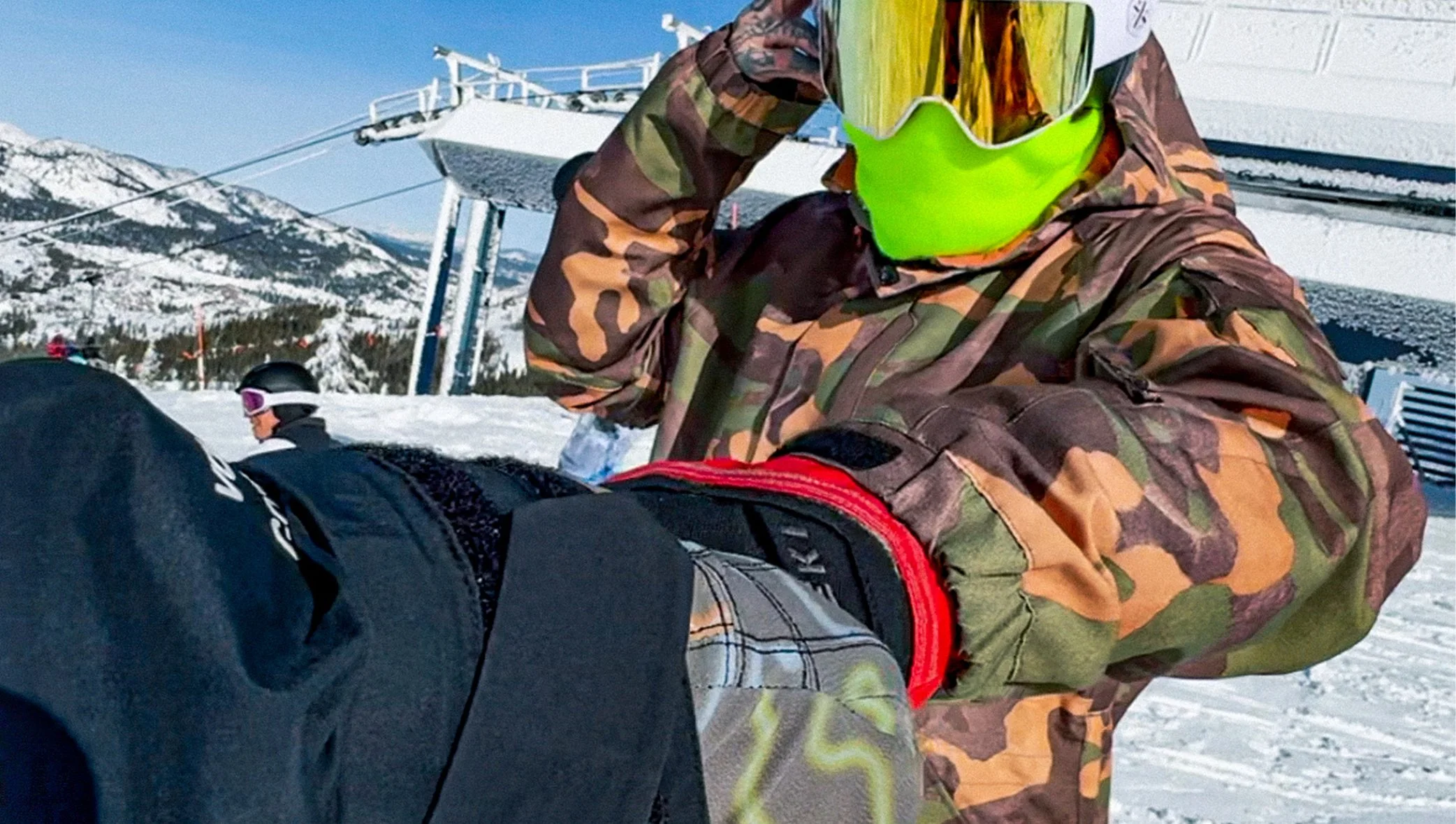 A person in camouflage jacket holding a snowboard, with other snowboarders and snowy mountains in the background at a ski resort.