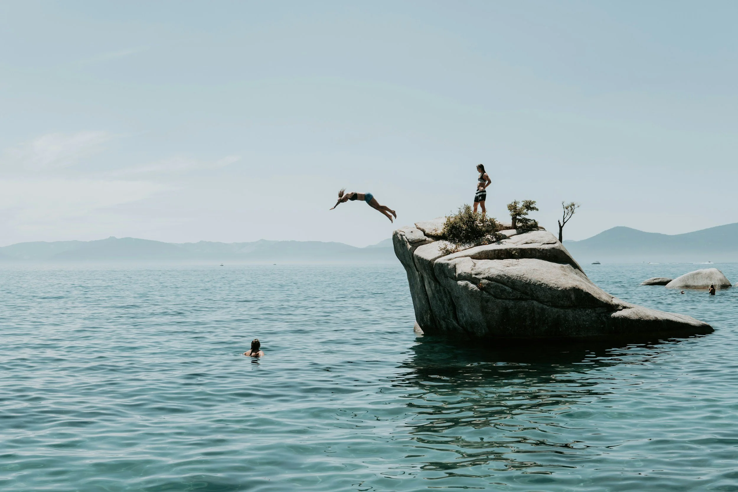 Two people on a large rock near a body of water; one person is jumping off into the water while the other stands on the rock watching, with a few trees on the rock and distant mountains in the background.