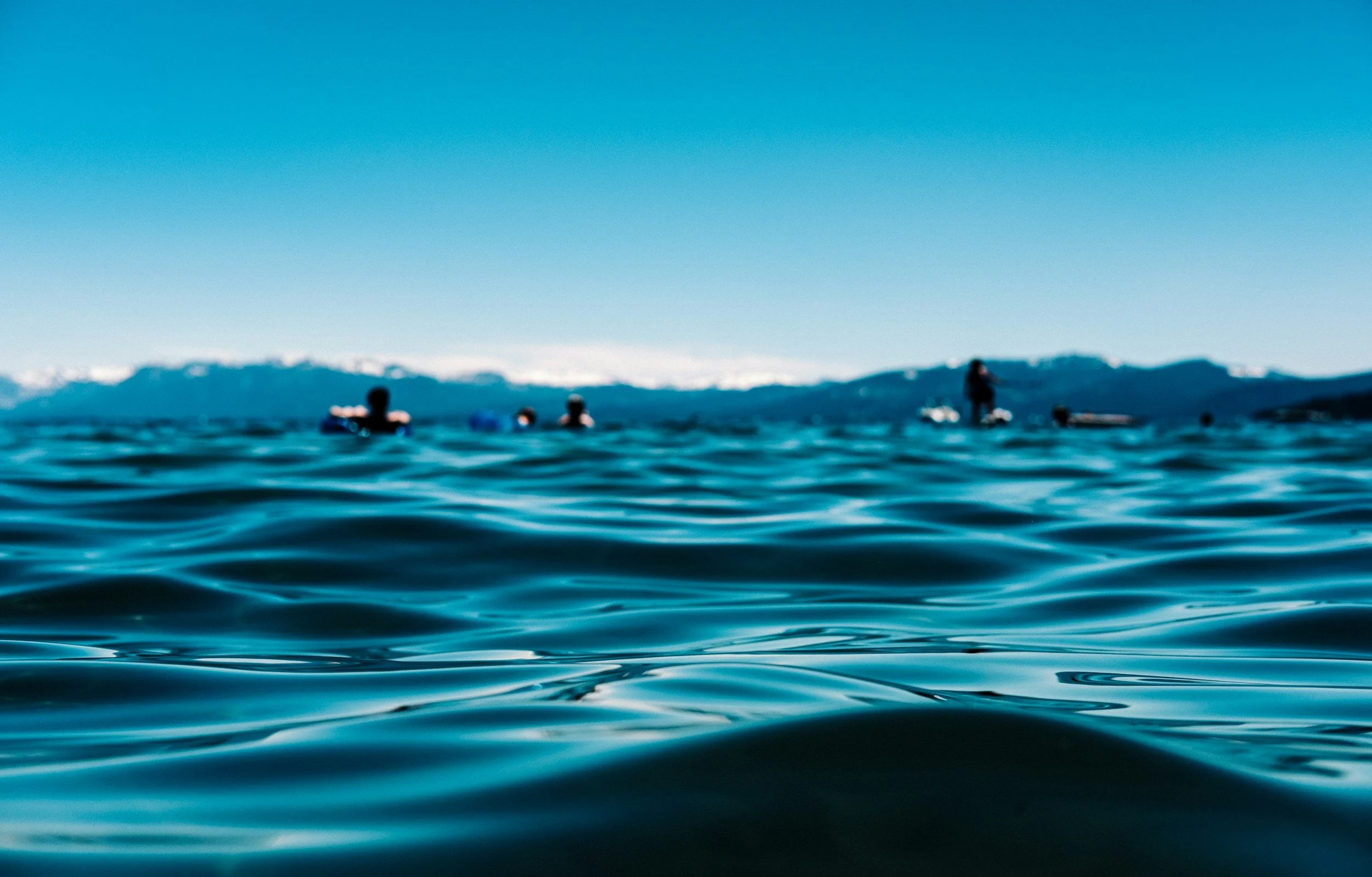 People swimming in an ocean with mountain range in the background under clear blue sky.