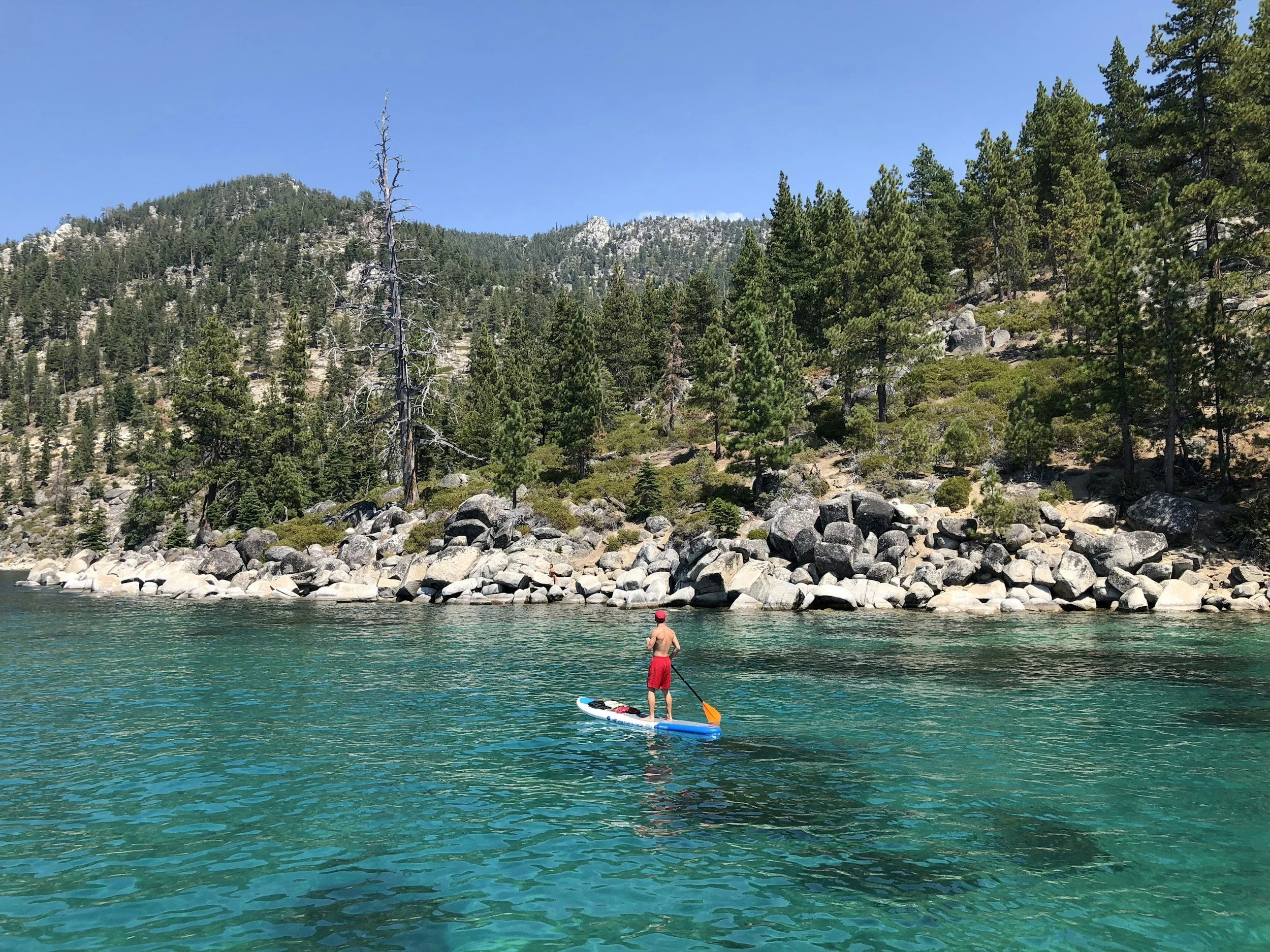 A man paddleboarding on a clear, turquoise lake with a rocky shoreline and a forested mountain in the background on a sunny day.