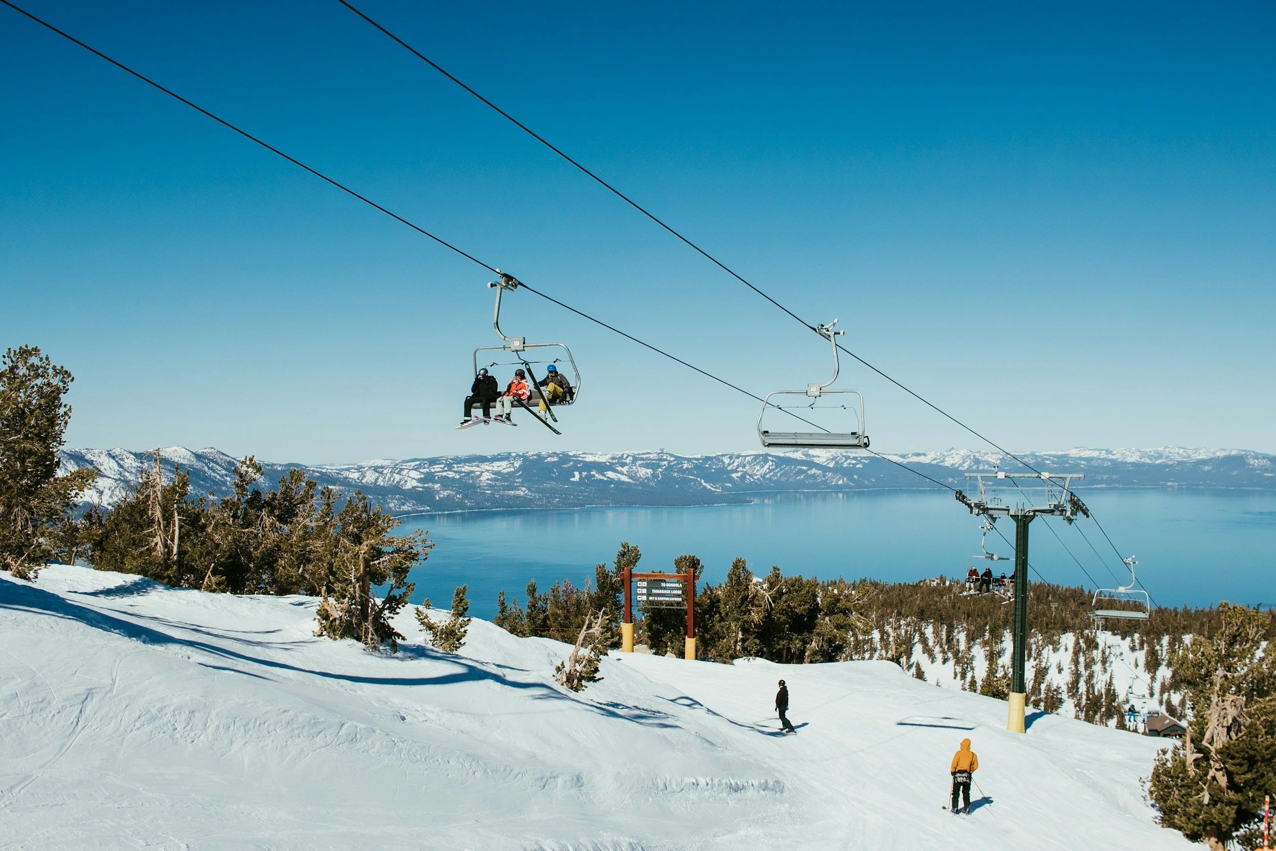 People riding ski lift over snowy slope with trees and a mountain lake in the background under clear blue sky.