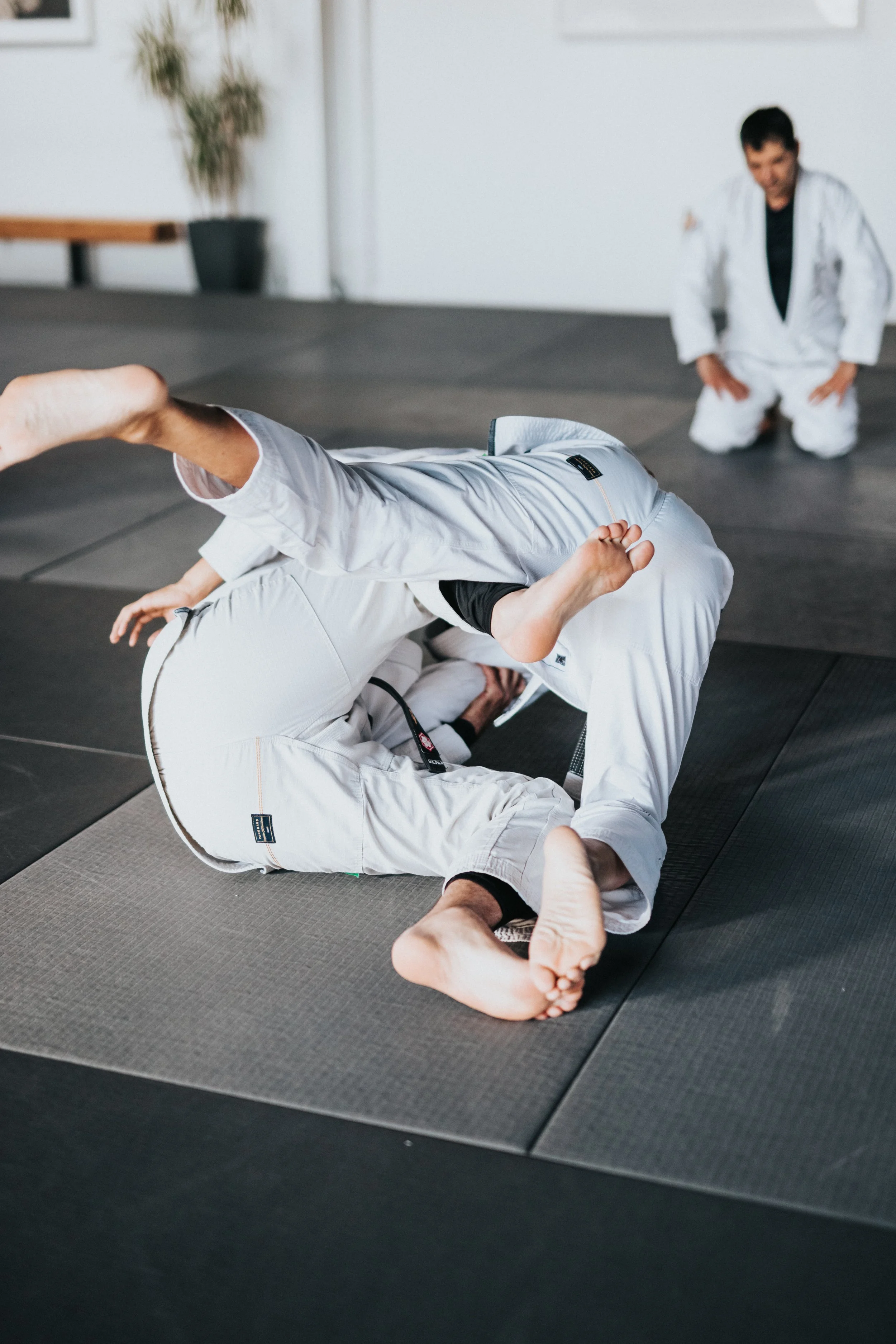 Two people practicing Brazilian Jiu-Jitsu on the mat, one person in a white gi executing a submission move on the other in a black gi, with an instructor observing in the background.