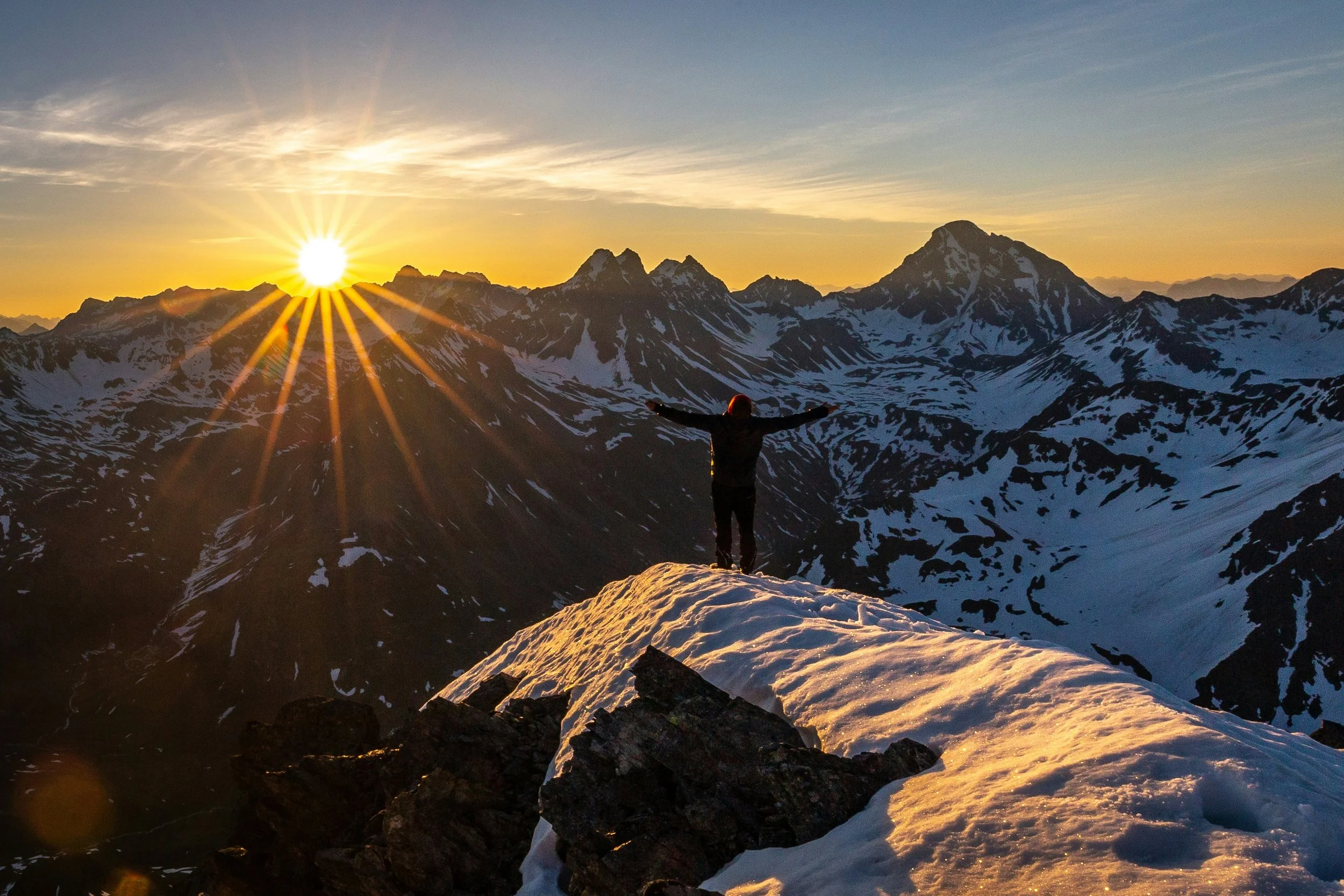 Person standing on a snow-covered mountain peak during sunrise with sunbeams and distant mountain range.