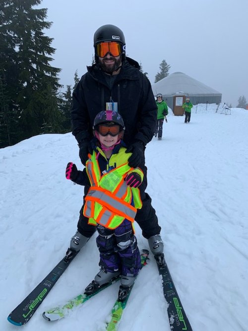 A young girl on skis and wearing ski pants, helmet and yellow and orange reflective vest stands in front of her father, also on skis.