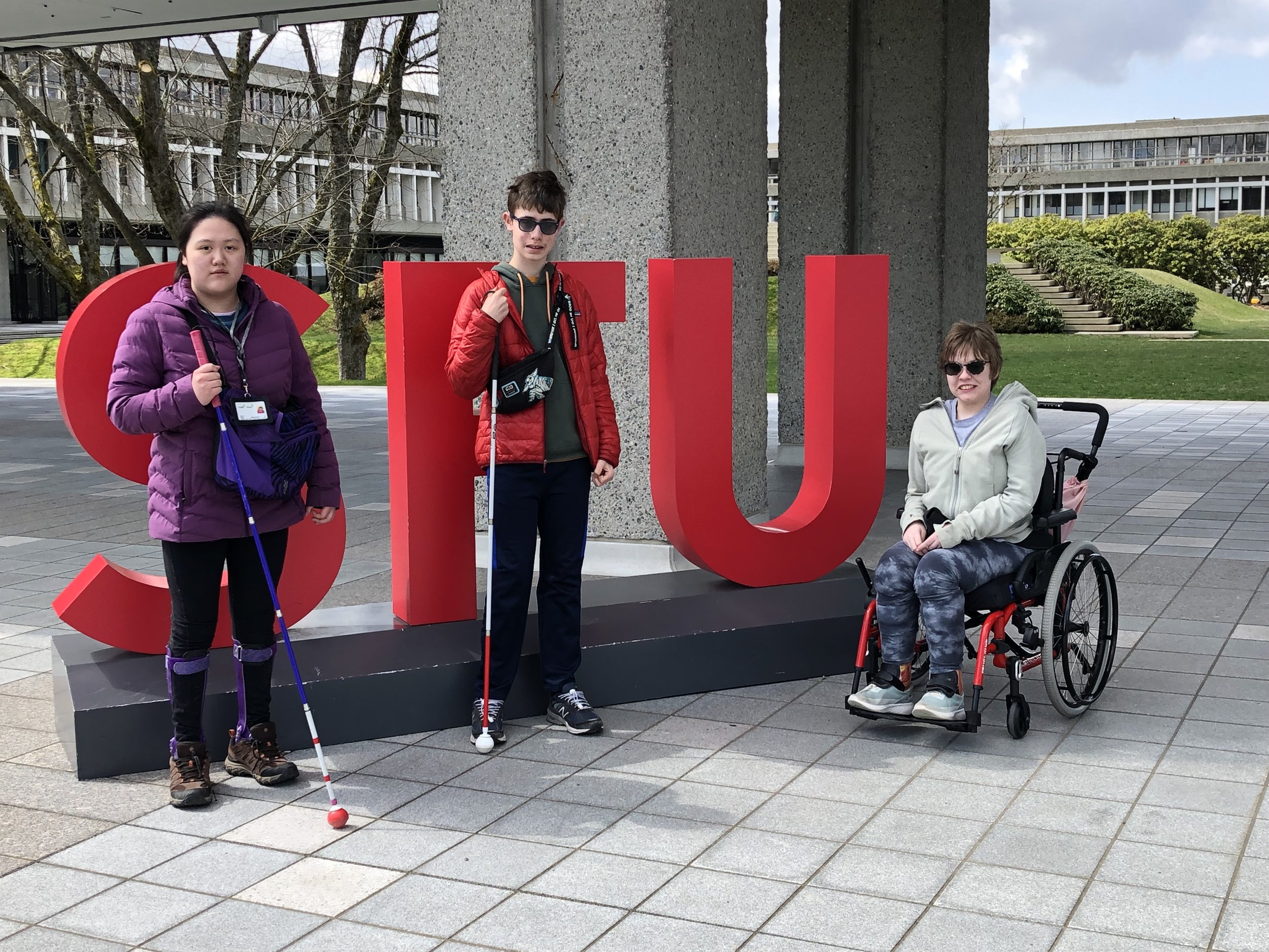 Three teens stand in front of three large letters that spell "SFU" on the grounds of Simon Fraser University. Two hold their mobility canes while the third sits in a wheelchair.
