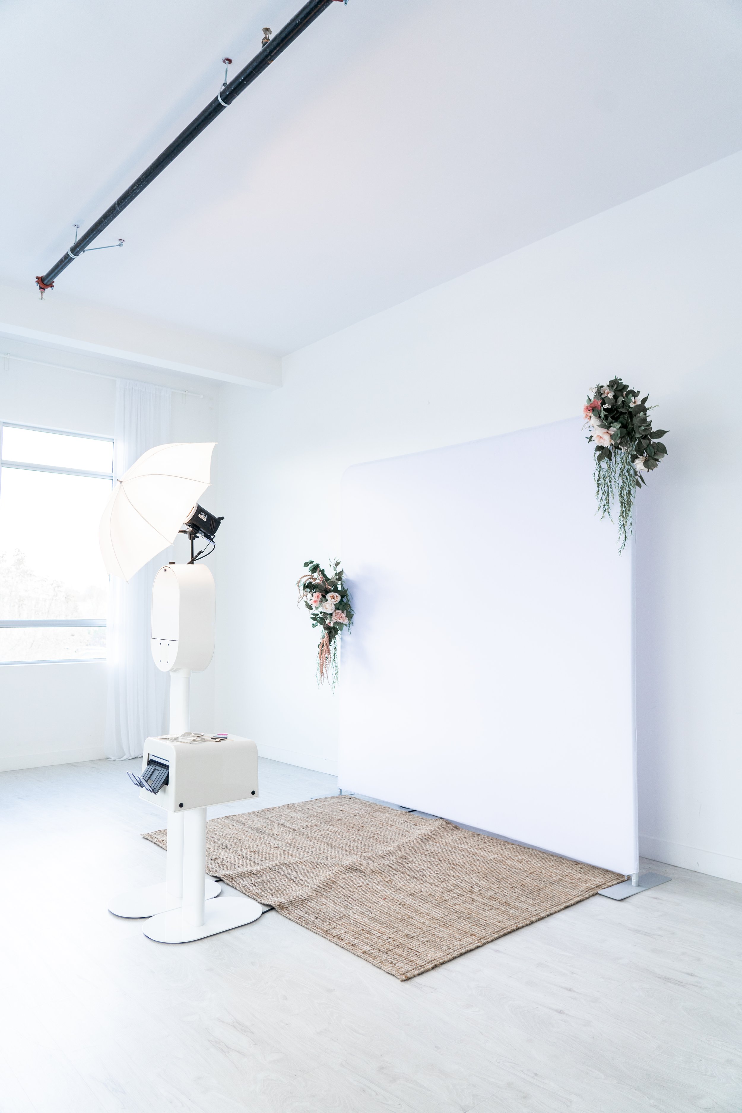 Photo booth setup in a bright room with white backdrop, two floral decorations, a photo machine, and a lighting umbrella. There is a textured rug on the floor.