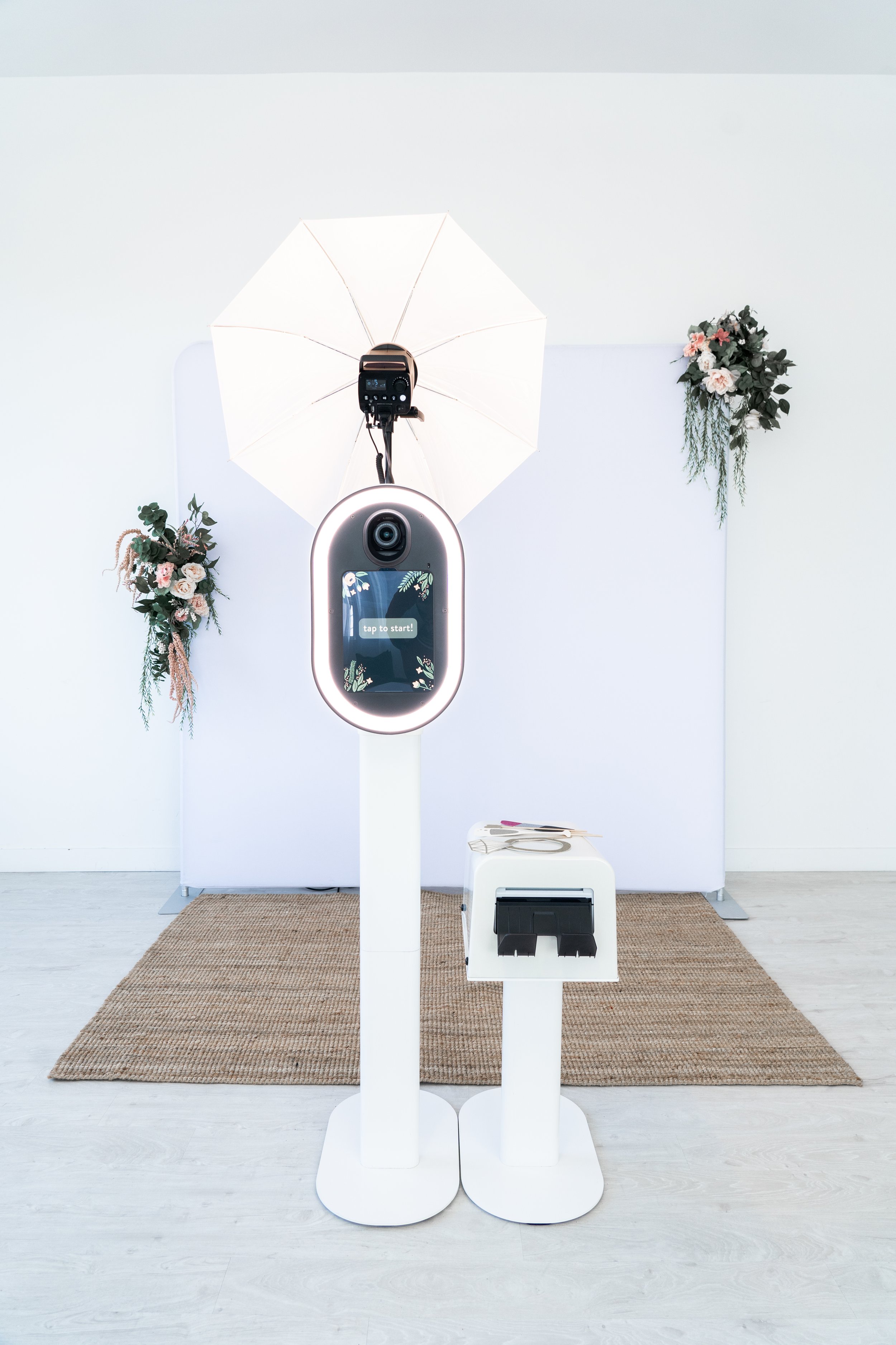Photo booth setup with a camera, umbrella light, tablet interface, and printer, placed on a rug with floral decorations in the background.