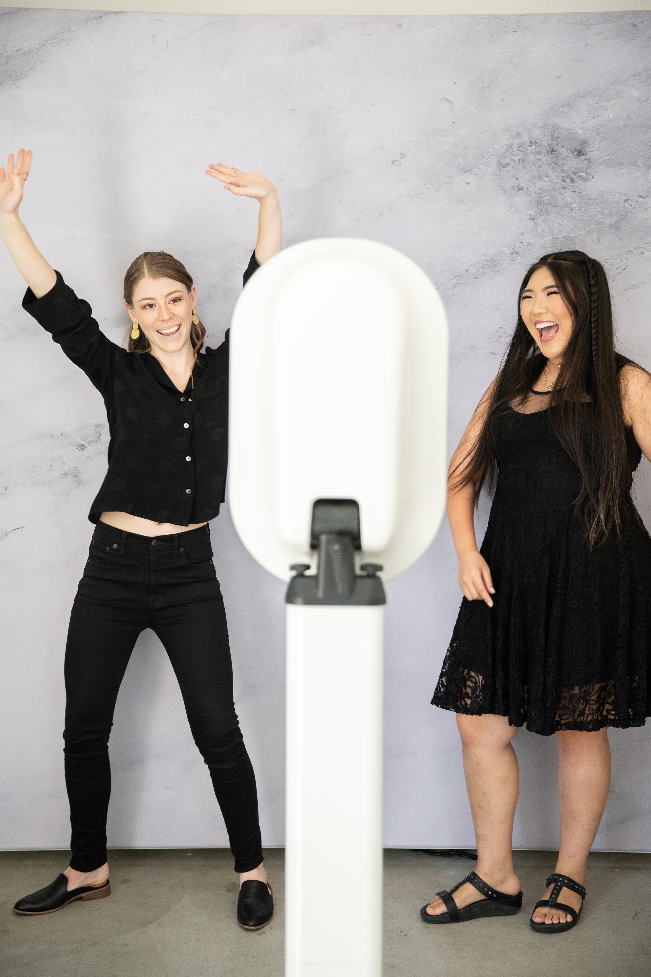 Two women posing joyfully for a camera in front of a gray backdrop. One woman raises her arms while the other laughs, both wearing black outfits.