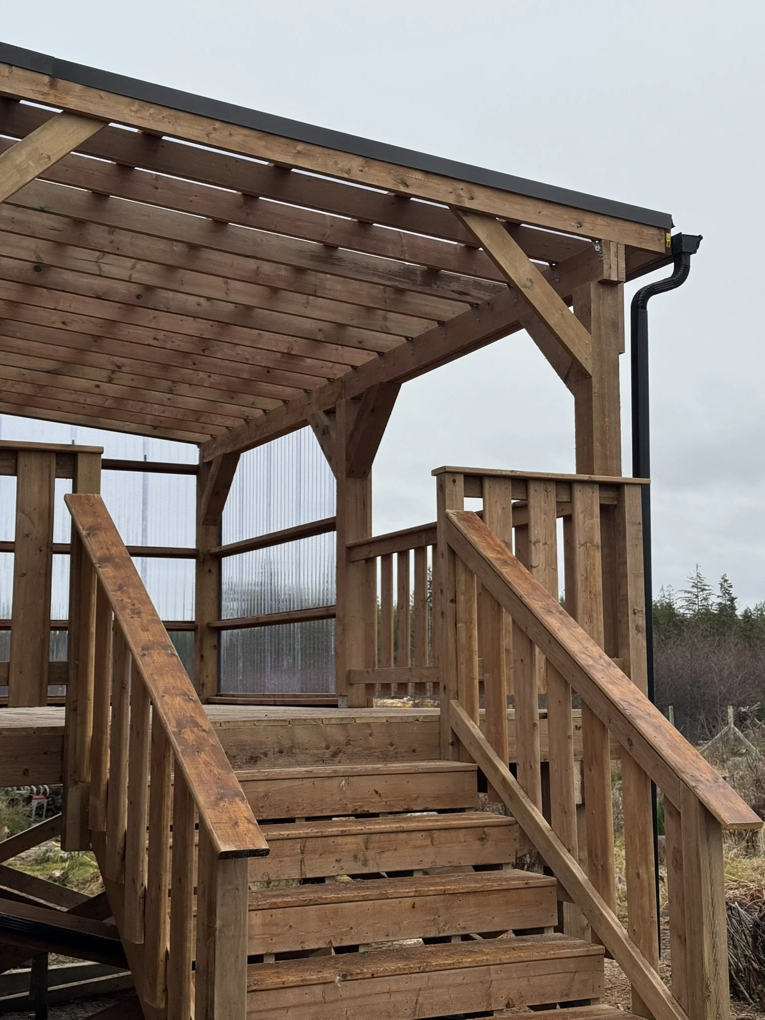 A wooden deck with stairs and railings, showing some support beams and a black rain gutter on the corner, against a cloudy sky and trees in the background.