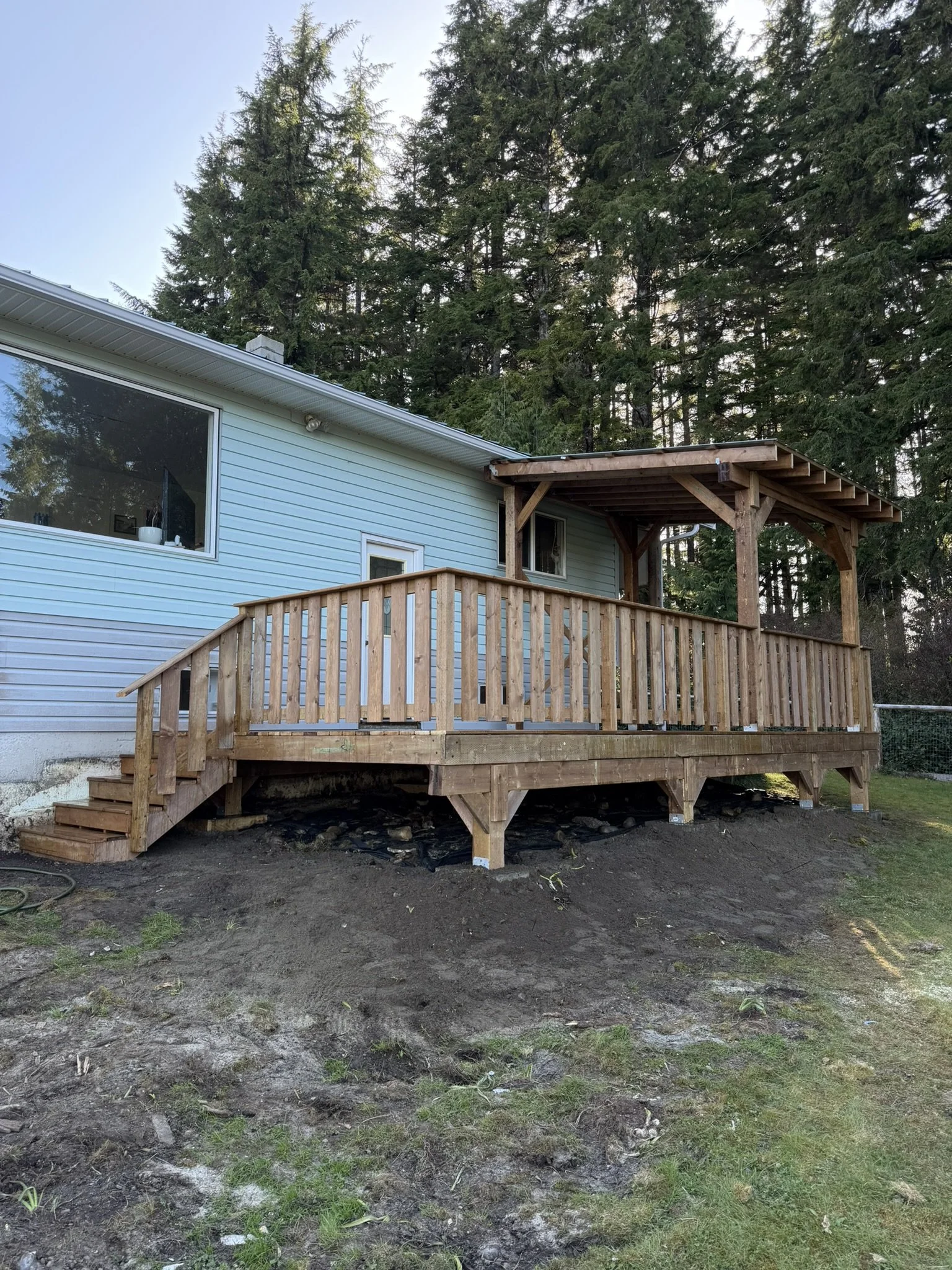 A house with light blue siding and a large window on the left side and a wooden deck with a roof extension on the right side, surrounded by tall trees in the background, and a patch of dirt and grass in the foreground.
