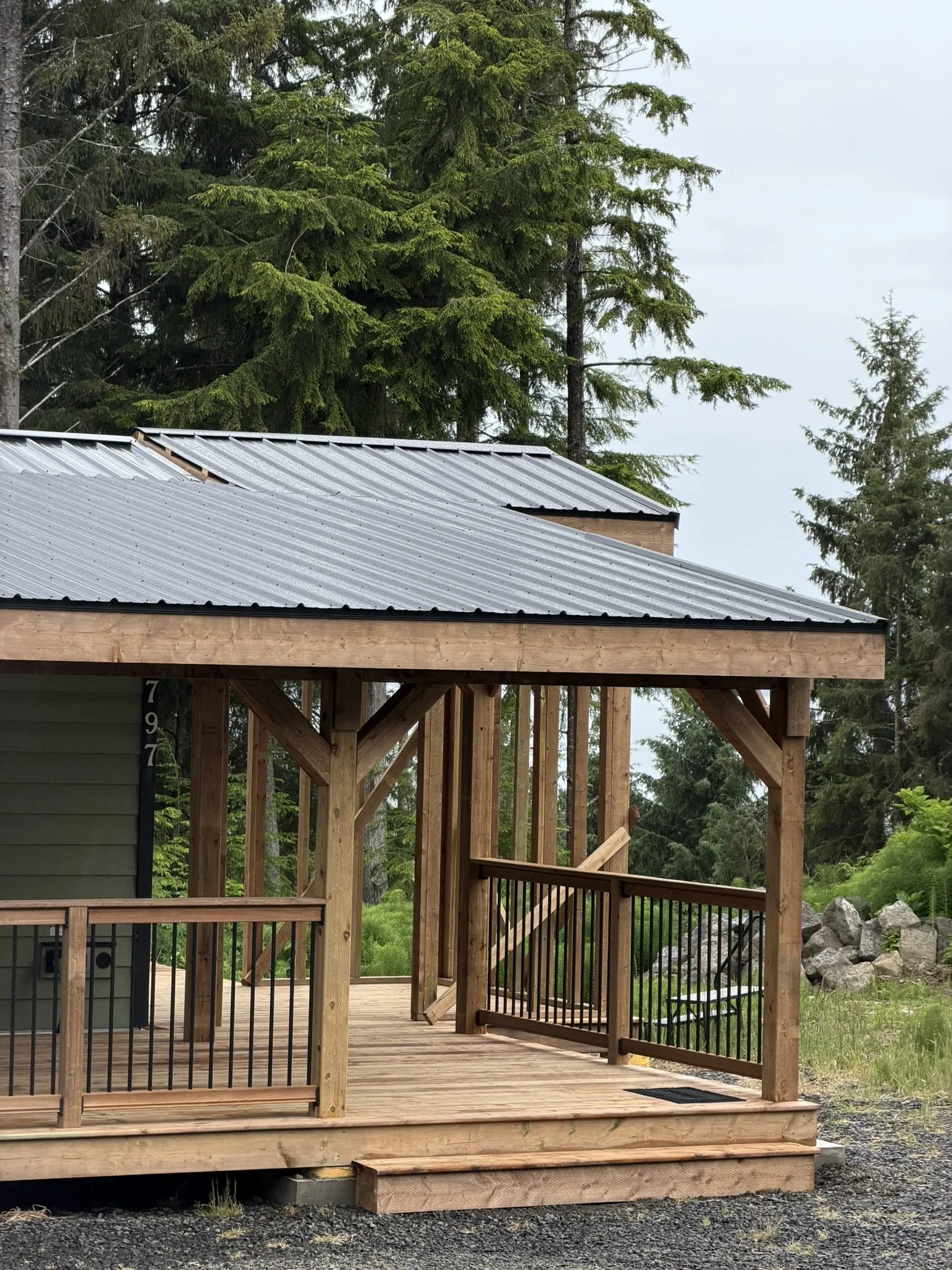 A wooden deck under construction with vertical beams and a corrugated metal roof, surrounded by green trees and rocks in the background.