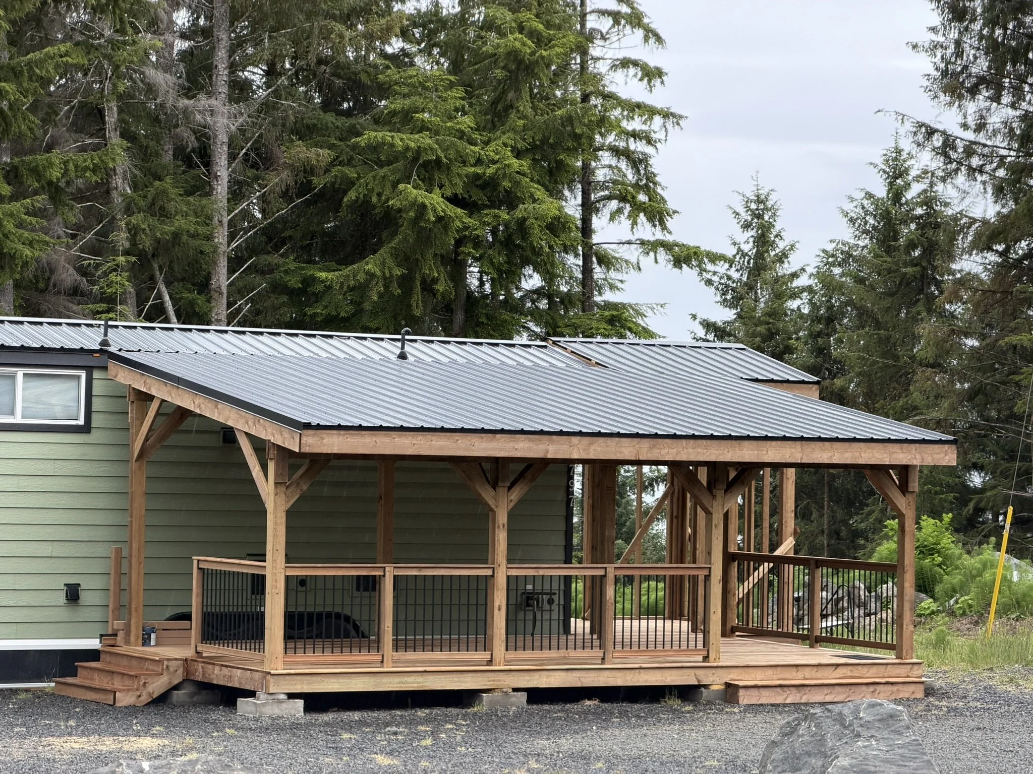 A house with light green exterior and black roof, under construction with a wooden deck and railing, set against a background of tall green pine trees and a cloudy sky.