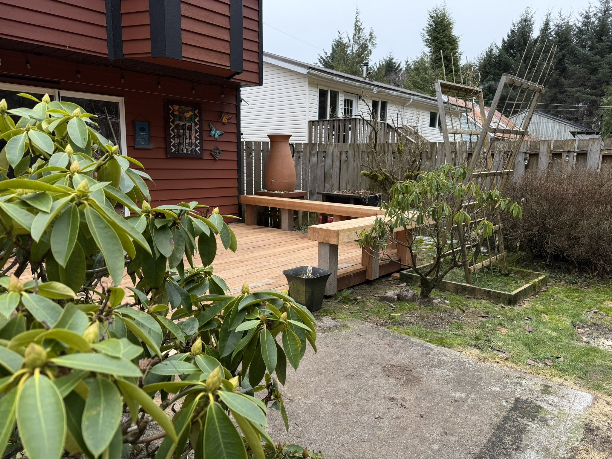 A backyard scene showing a wooden deck, a variety of green bushes, a white house with a deck railing, a tall red house with a decorated window, and an outdoor wooden garden structure with ladders. In the background, there are trees under a mostly clo