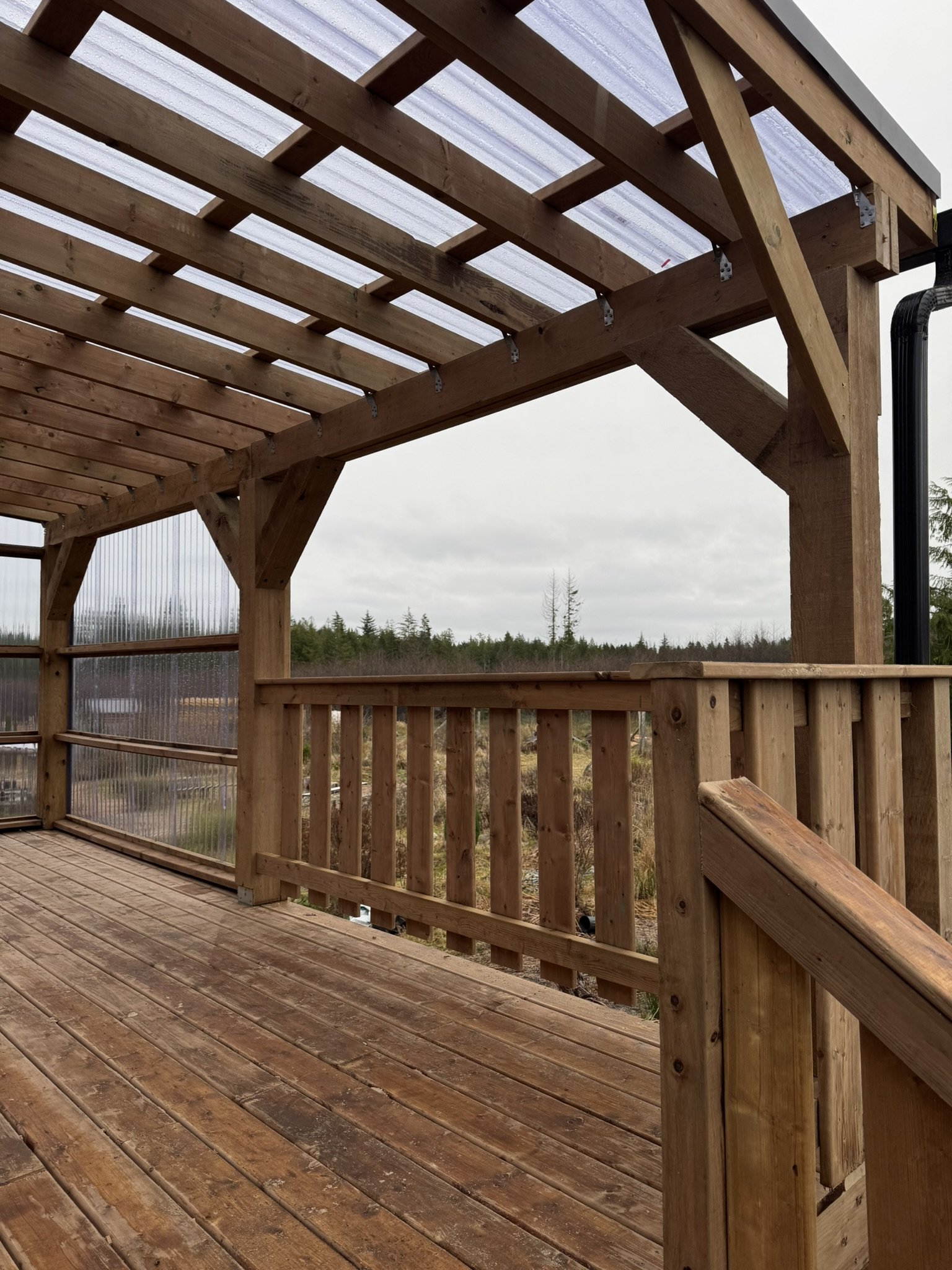 Wooden deck with railing and partial roof, overlooking a landscape with trees and cloudy sky.