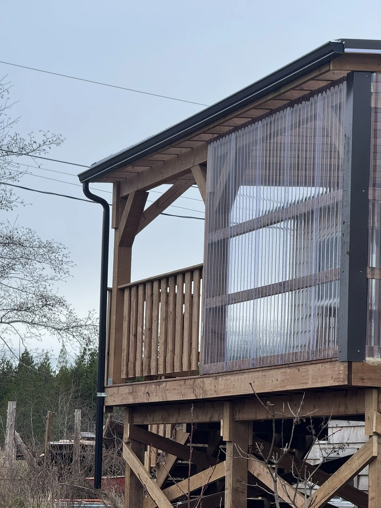A partially constructed wooden deck with a clear plastic privacy screen and a black metal roof overhang, supported by wooden beams, built on the second floor of a house. Trees and utility wires are visible in the background.