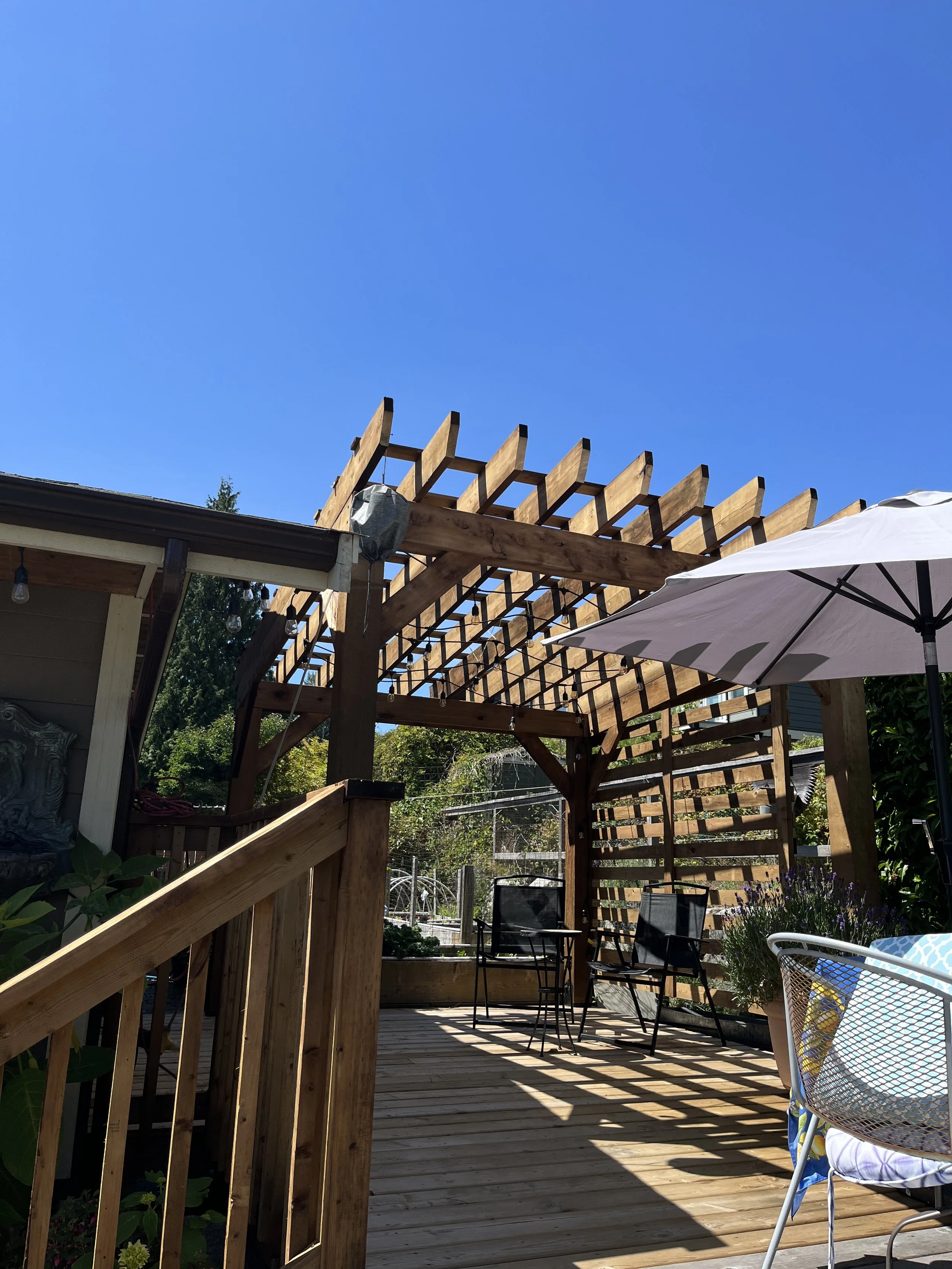 A wooden deck patio with a pergola, outdoor table, and chairs, a white patio umbrella, and lush green trees and plants, under a clear blue sky.