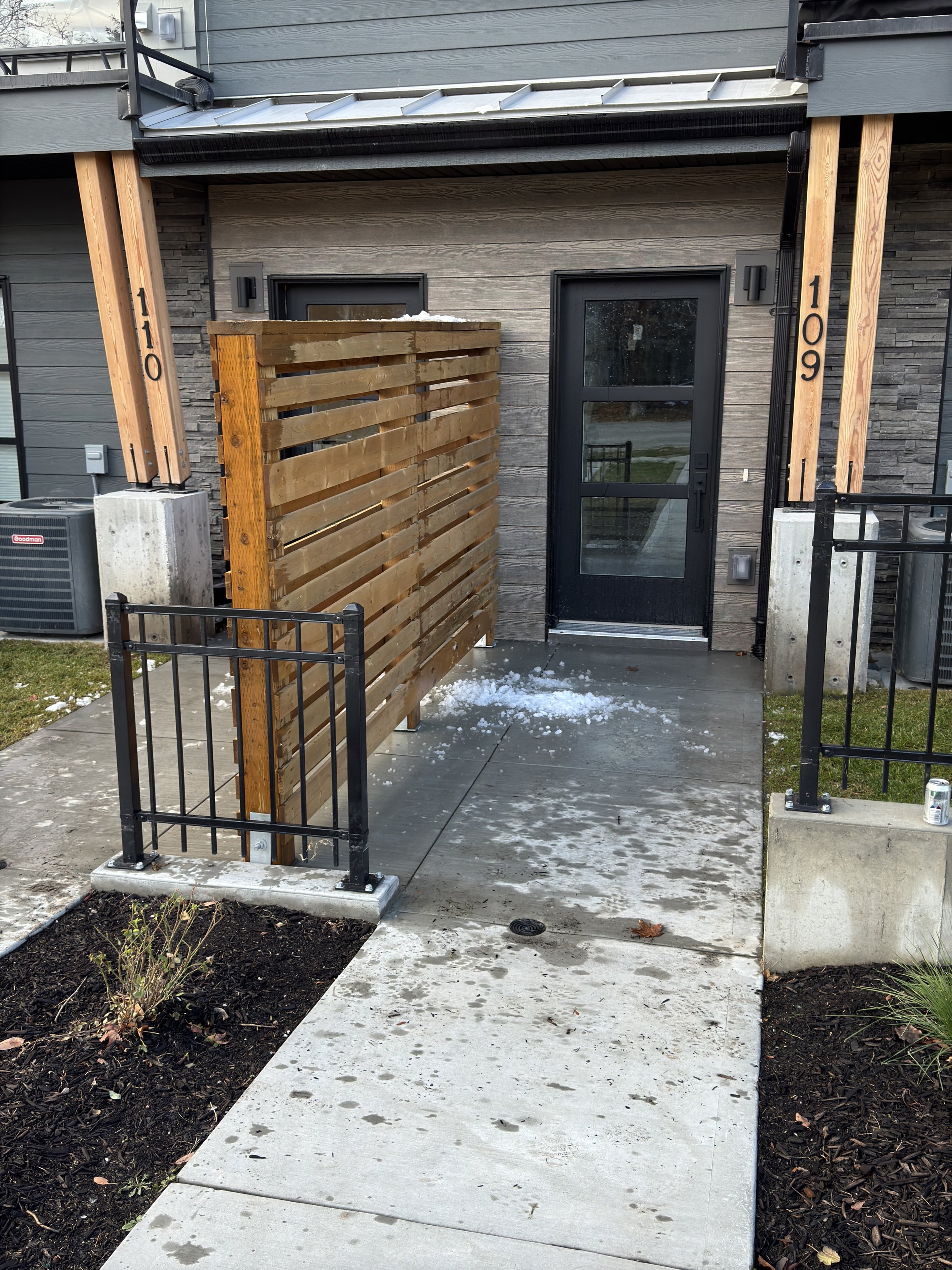 Front door of a modern apartment with a small fenced area, wooden privacy screen, concrete walkway, plants, and snow on the ground.