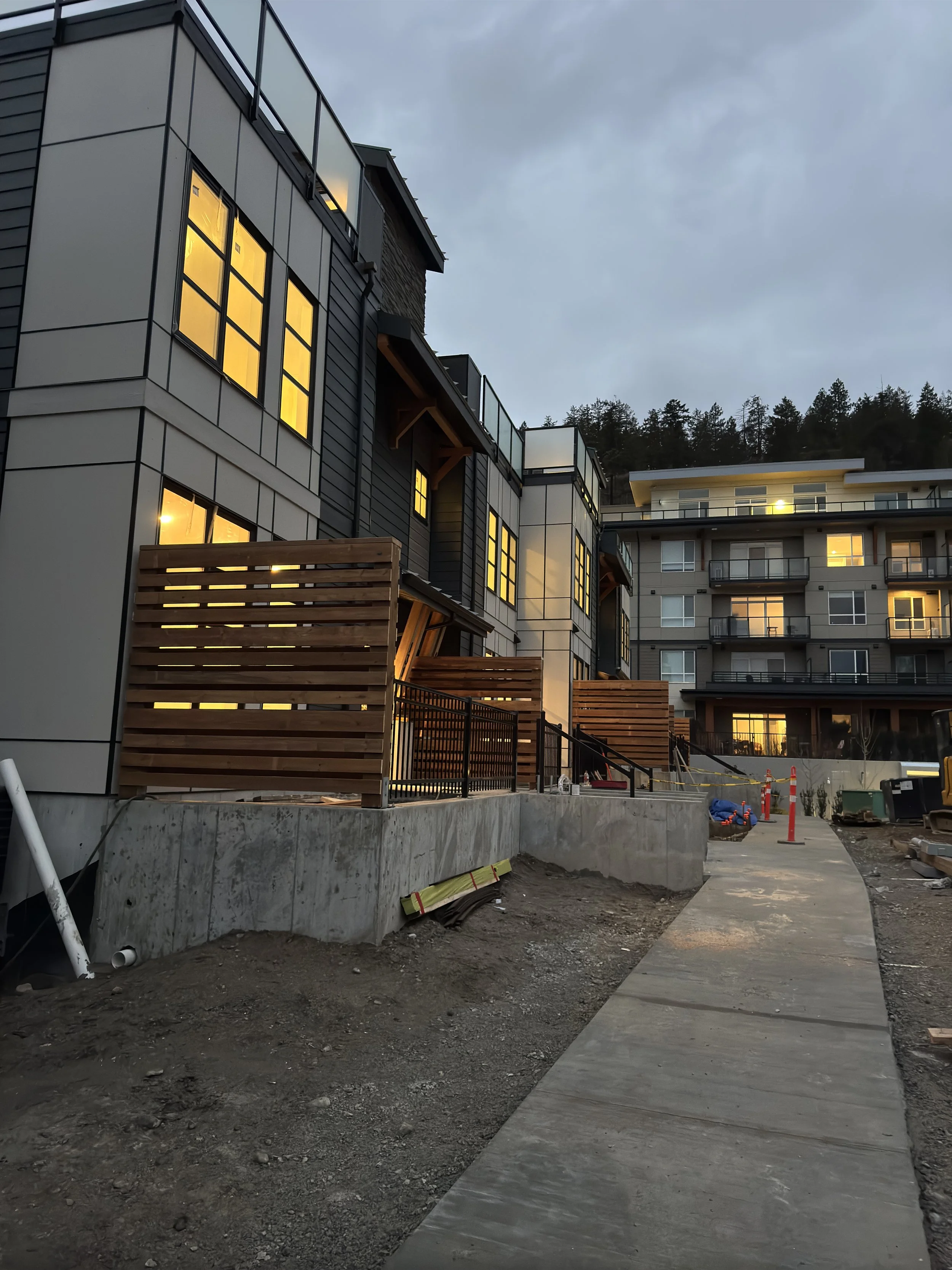 Construction site with newly built apartment buildings, some windows lit, wooden fencing, and a concrete sidewalk under cloudy sky.