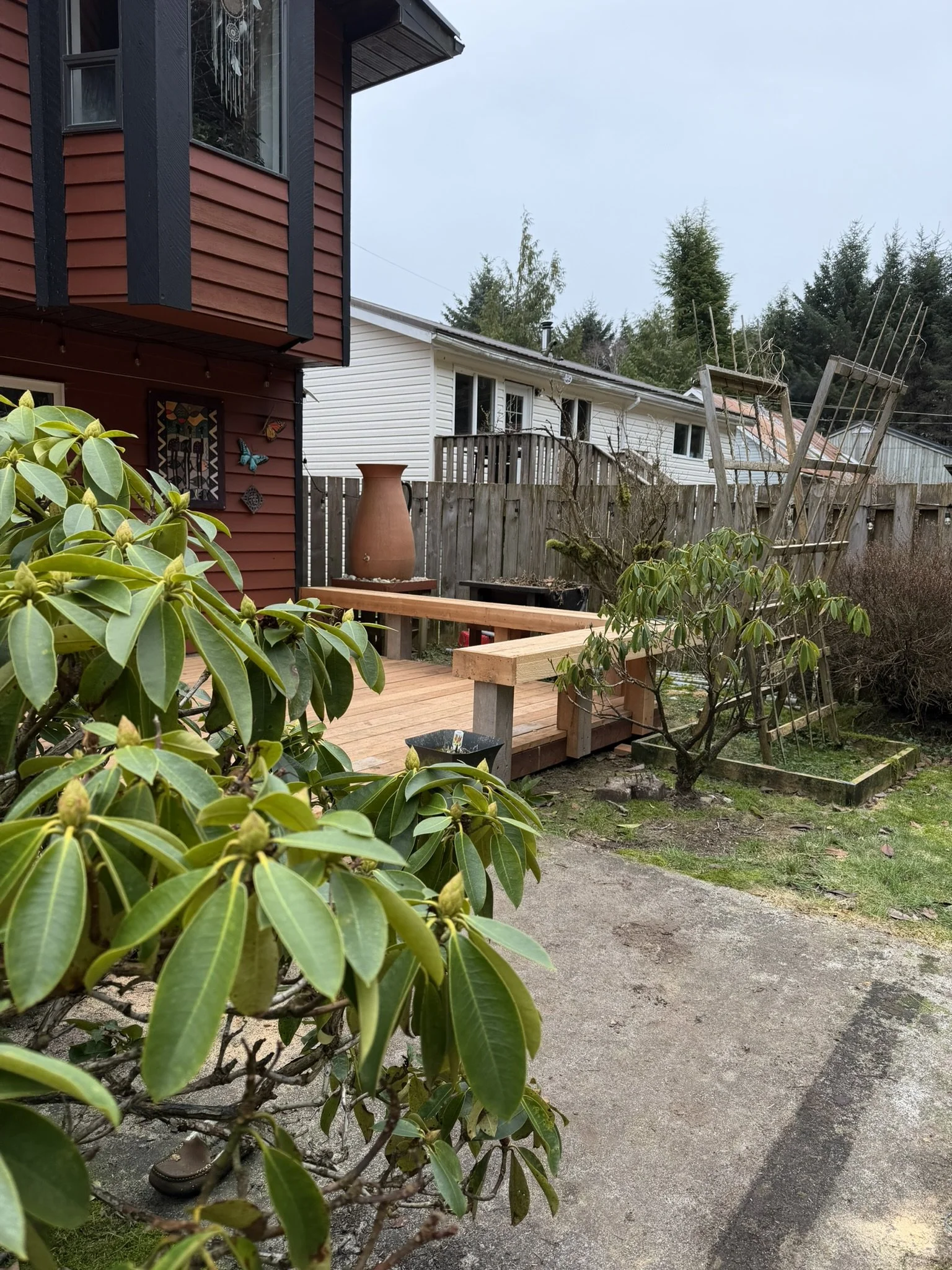 A backyard scene with a deck, green shrubbery, a wooden ladder leaning against a fence, and a white house with a balcony in the background. There are trees and a cloudy sky overhead.