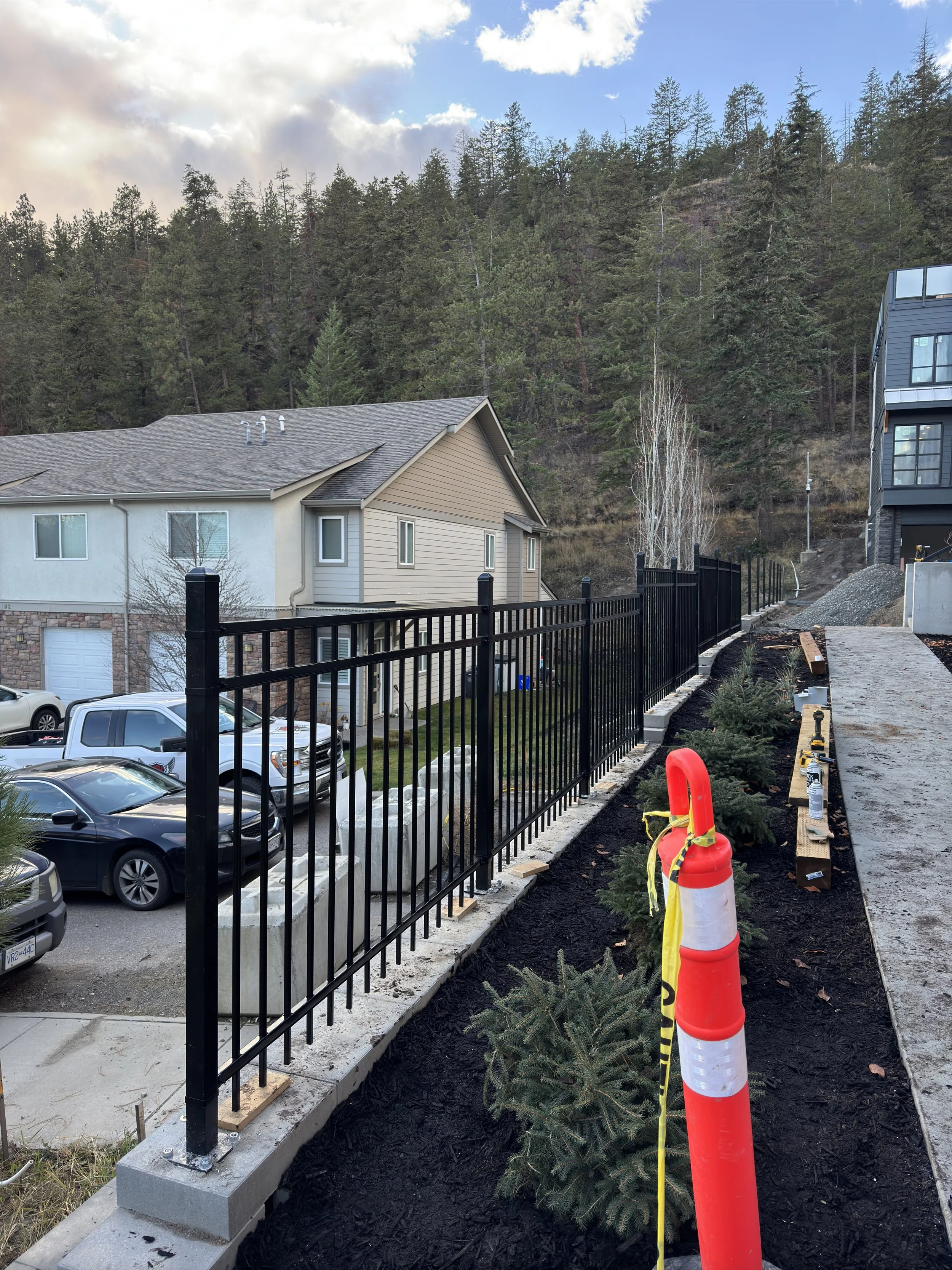 A black metal fence being installed along a concrete walkway, with construction tools and a safety cone in the foreground, residential houses behind, and a forested hillside in the background.