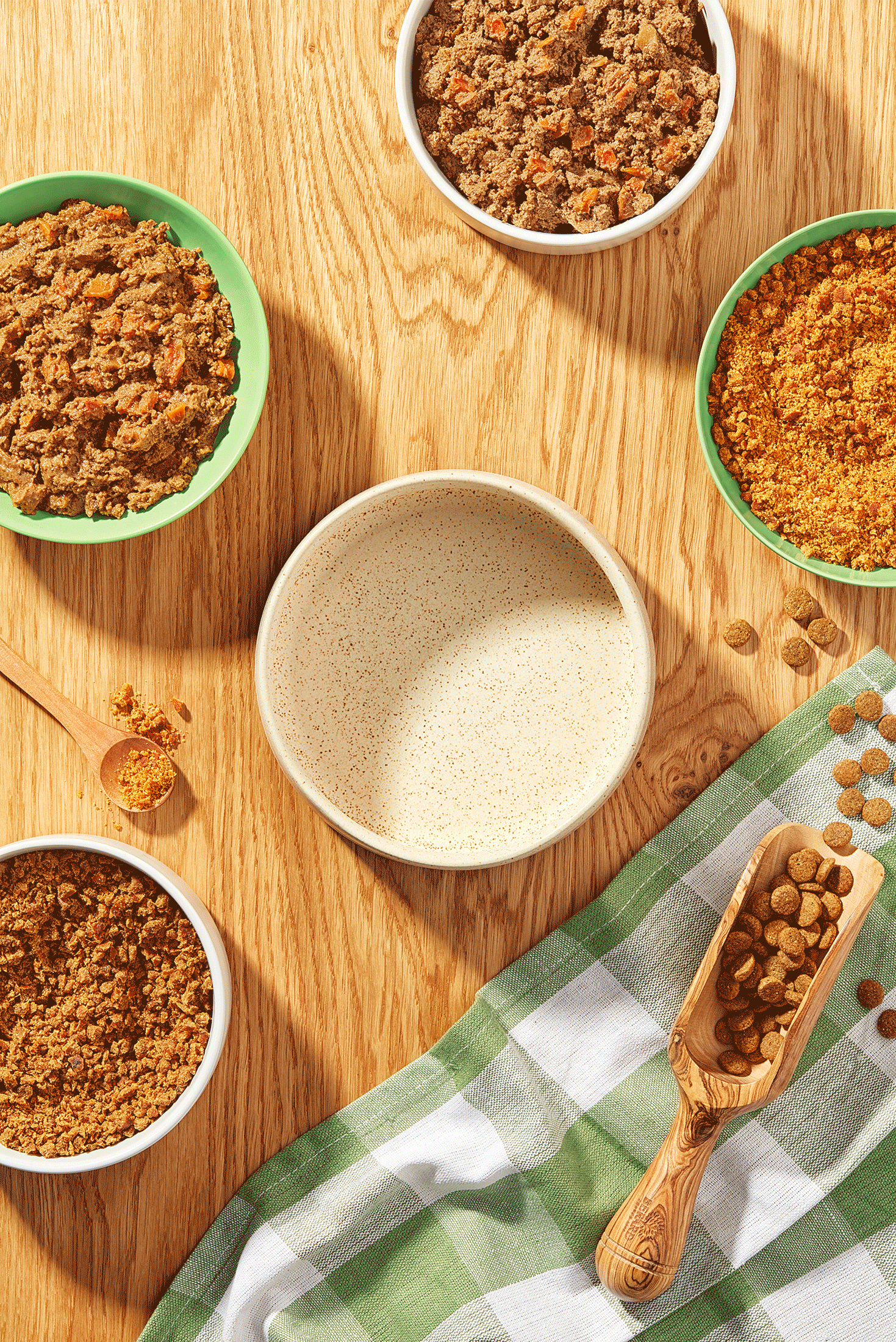 Variety of pet food in bowls and wooden scoop on a wooden table with checkered cloth.