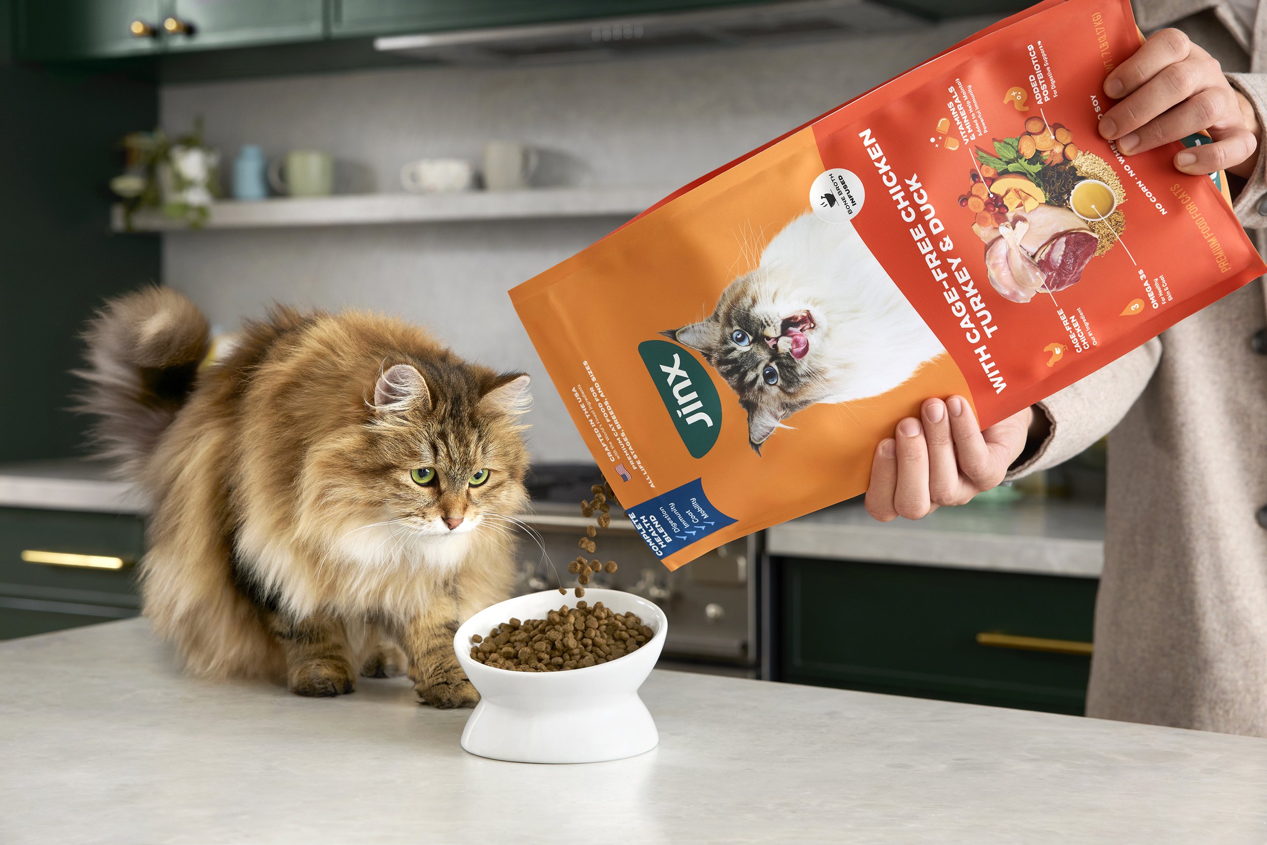 A person pouring dry cat food from a bright orange bag labeled 'Jinx' into a white bowl on a kitchen countertop, with a fluffy tabby cat watching nearby.
