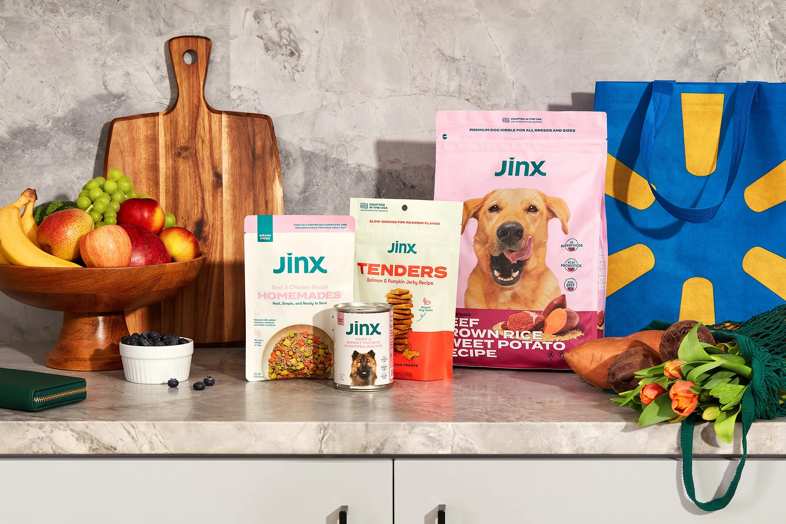 Variety of Jinx dog food products, including dry food and treats, displayed on a kitchen counter with fruits, sweet potatoes, a cutting board, a blue reusable bag with yellow design, and a small bouquet of orange flowers.