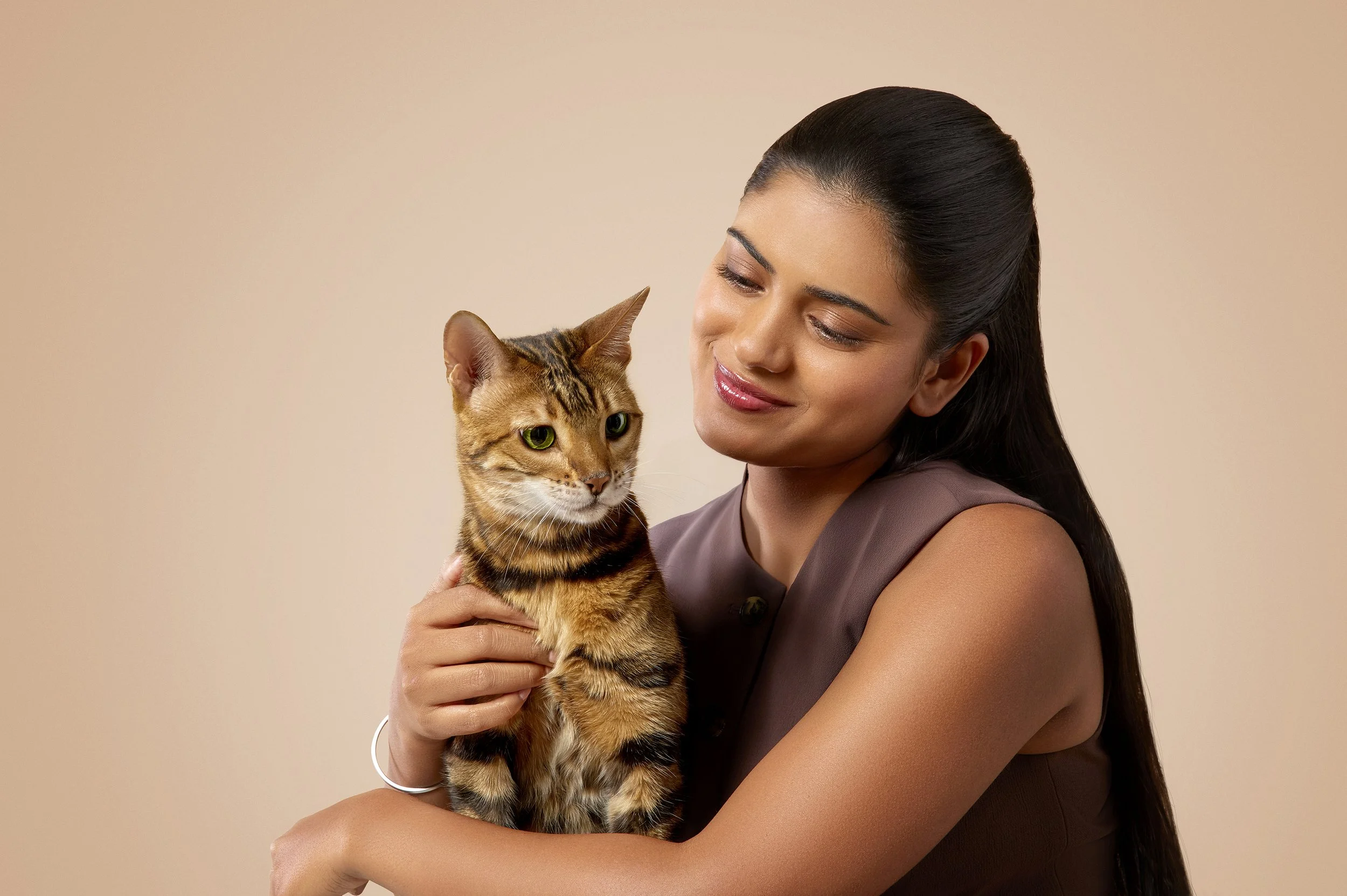 A woman with long dark hair holding a tabby cat close to her face, both looking at each other with a neutral expression, against a plain beige background.