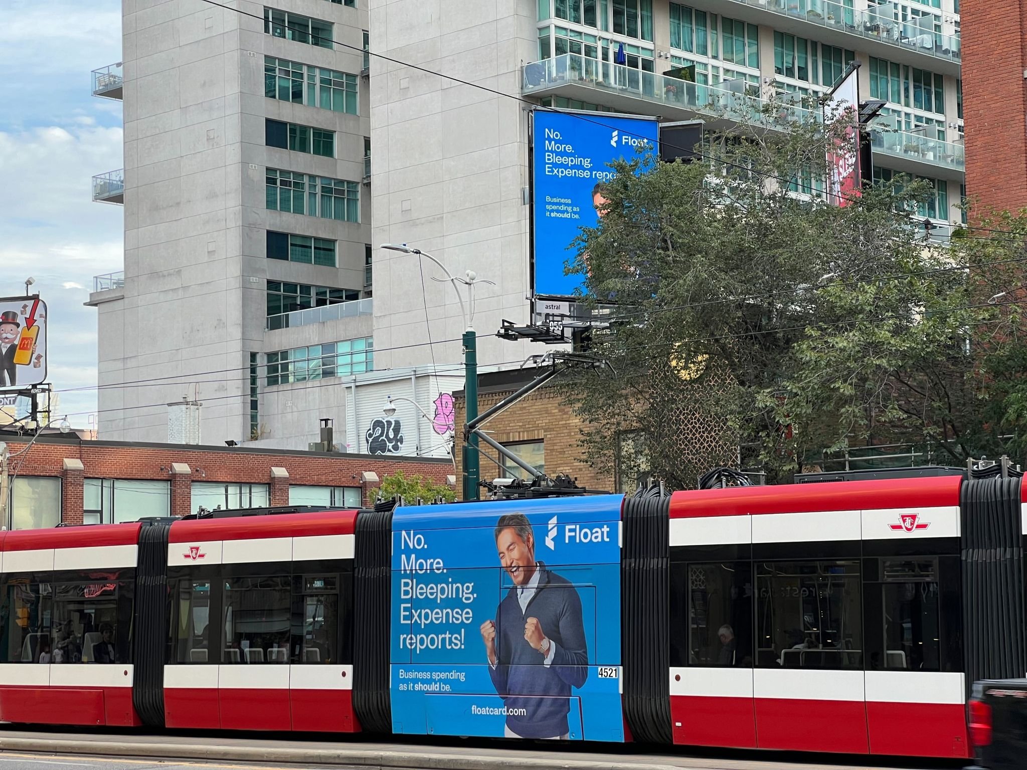Street scene with a tram featuring an advertisement for a business expense card service. The ad reads "No. More. Bleeping. Expense reports! Business spending as it should be. floatcard.com." A billboard in the background displays the same advertiseme